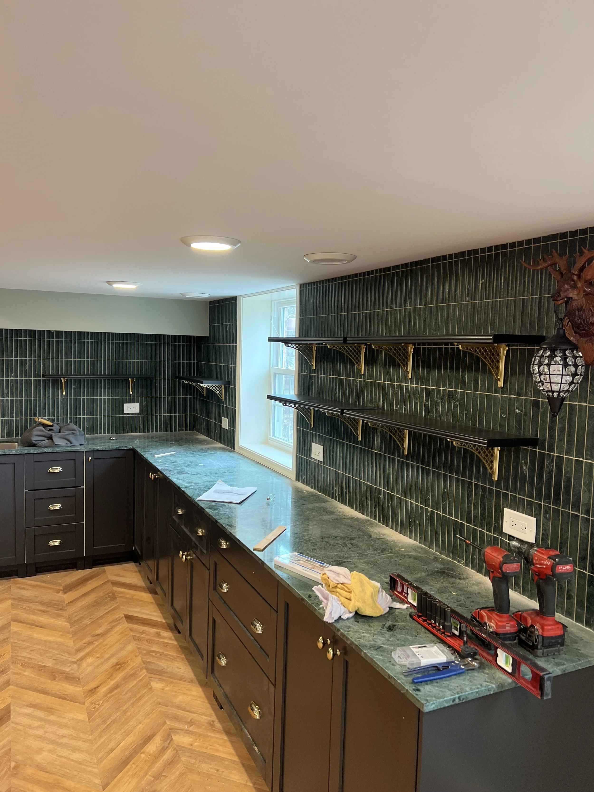 View of a kitchen under renovation, showing green marble countertops, black cabinets with gold handles, green tile backsplash, wall-mounted shelves, and power tools on the counter.