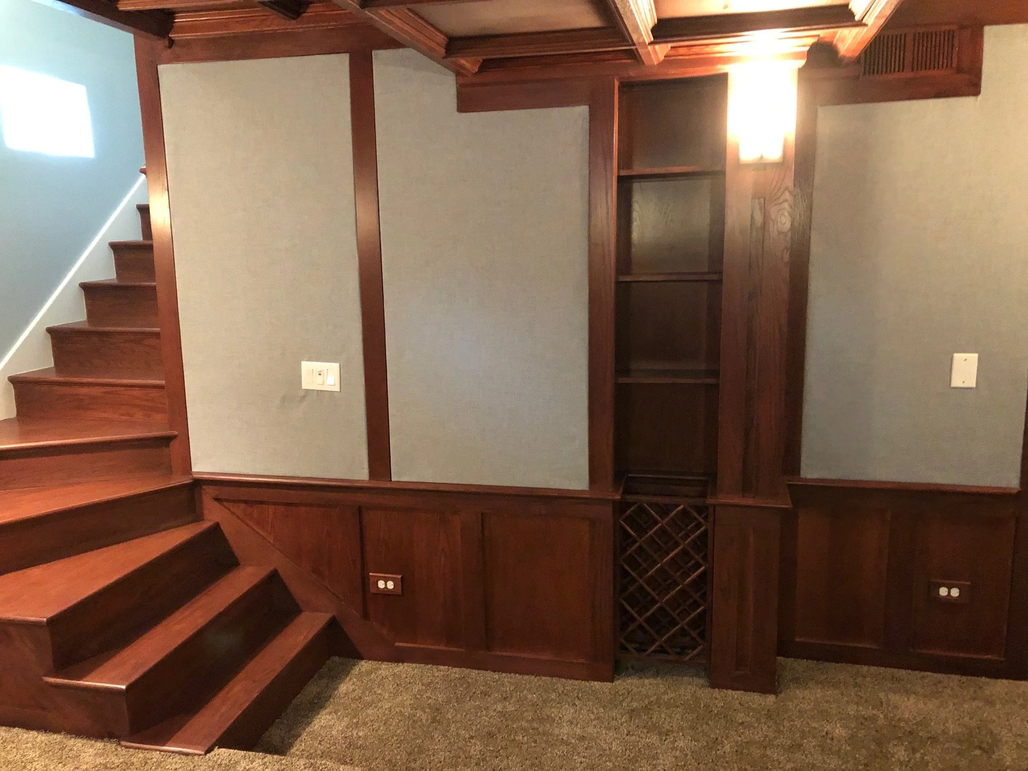 Interior of a room with a wood-paneled wall, built-in shelves, wine rack, a staircase with wooden steps, carpeted floor, and a wall-mounted light fixture.