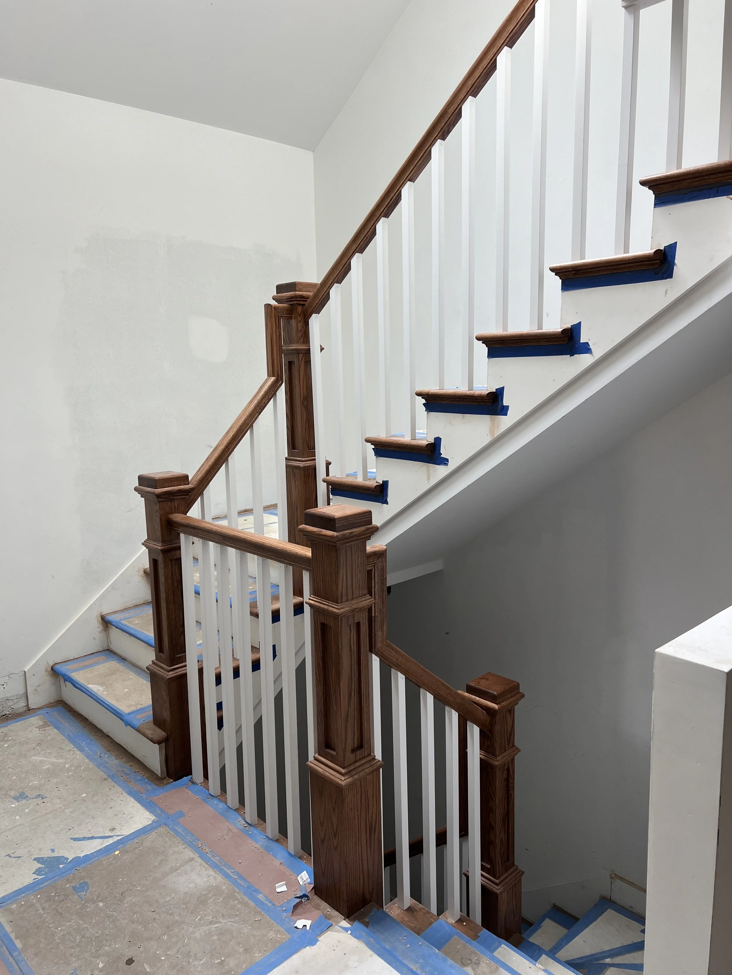 Interior view of a staircase under construction with new wooden handrails and white balusters, blue painter's tape on steps, and unfinished flooring.