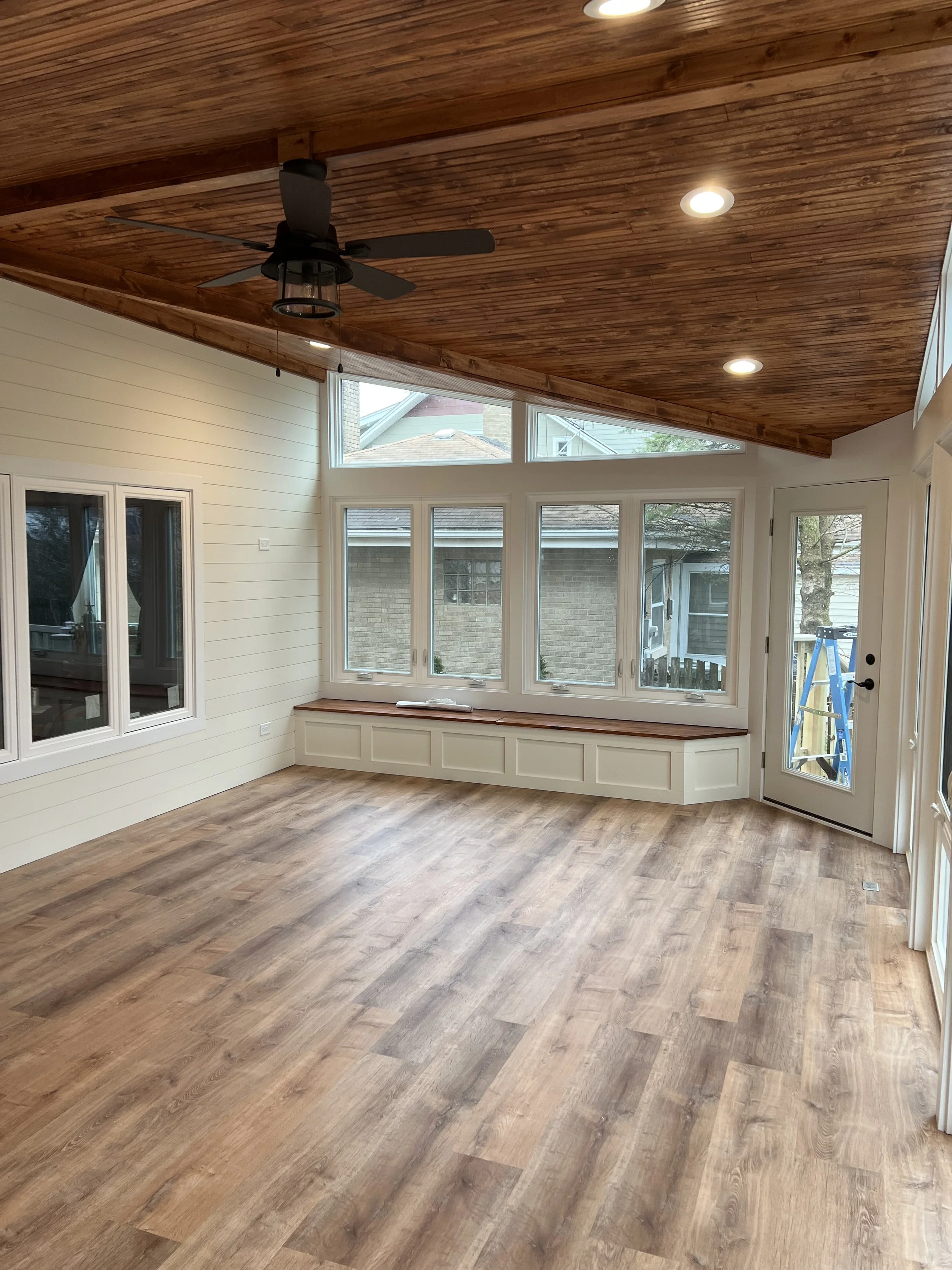 Empty sunroom with wood flooring, white paneled walls, large windows, a door with glass panel, and a wooden ceiling with a ceiling fan and recessed lighting.