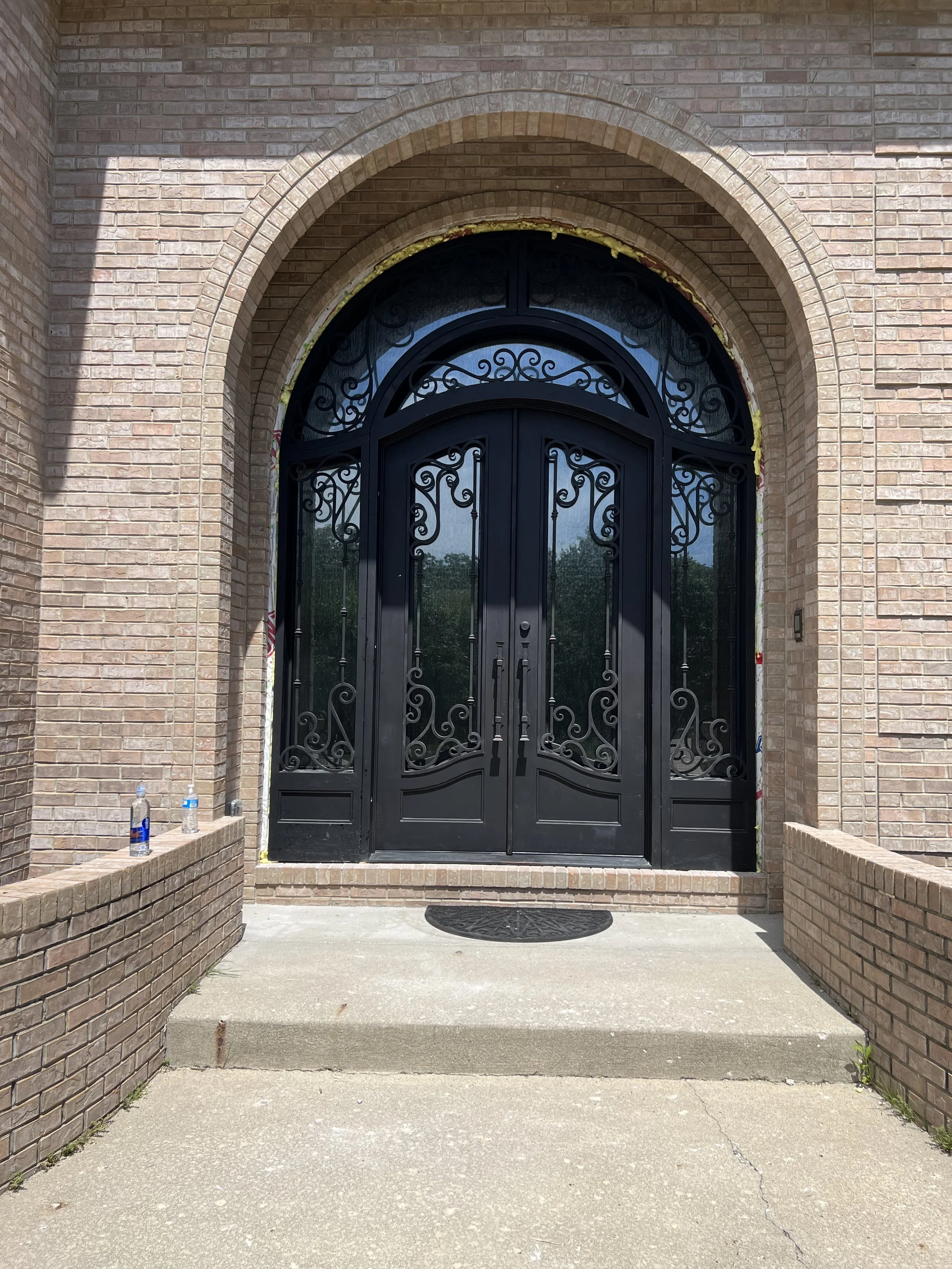 Black wrought iron double doors with decorative scrollwork, set in a brick archway, leading into a building. Two water bottles sit on the brick wall to the left of the steps. The area in front of the doors is paved, and a semi-circular doormat is on 