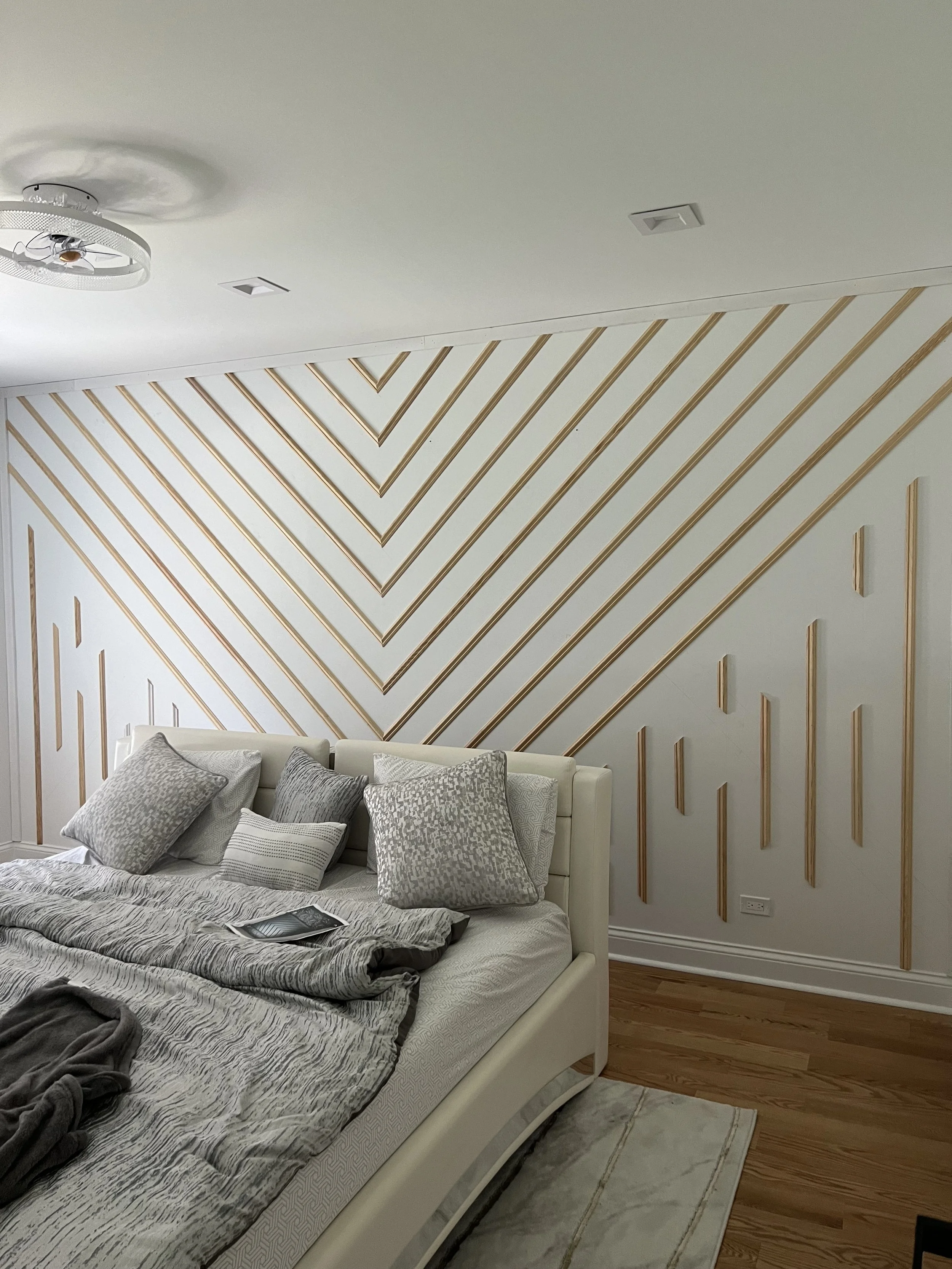 A bedroom featuring a white bed with various gray patterned pillows and a gray blanket, against a decorative wall with gold diagonal lines forming a geometric pattern.