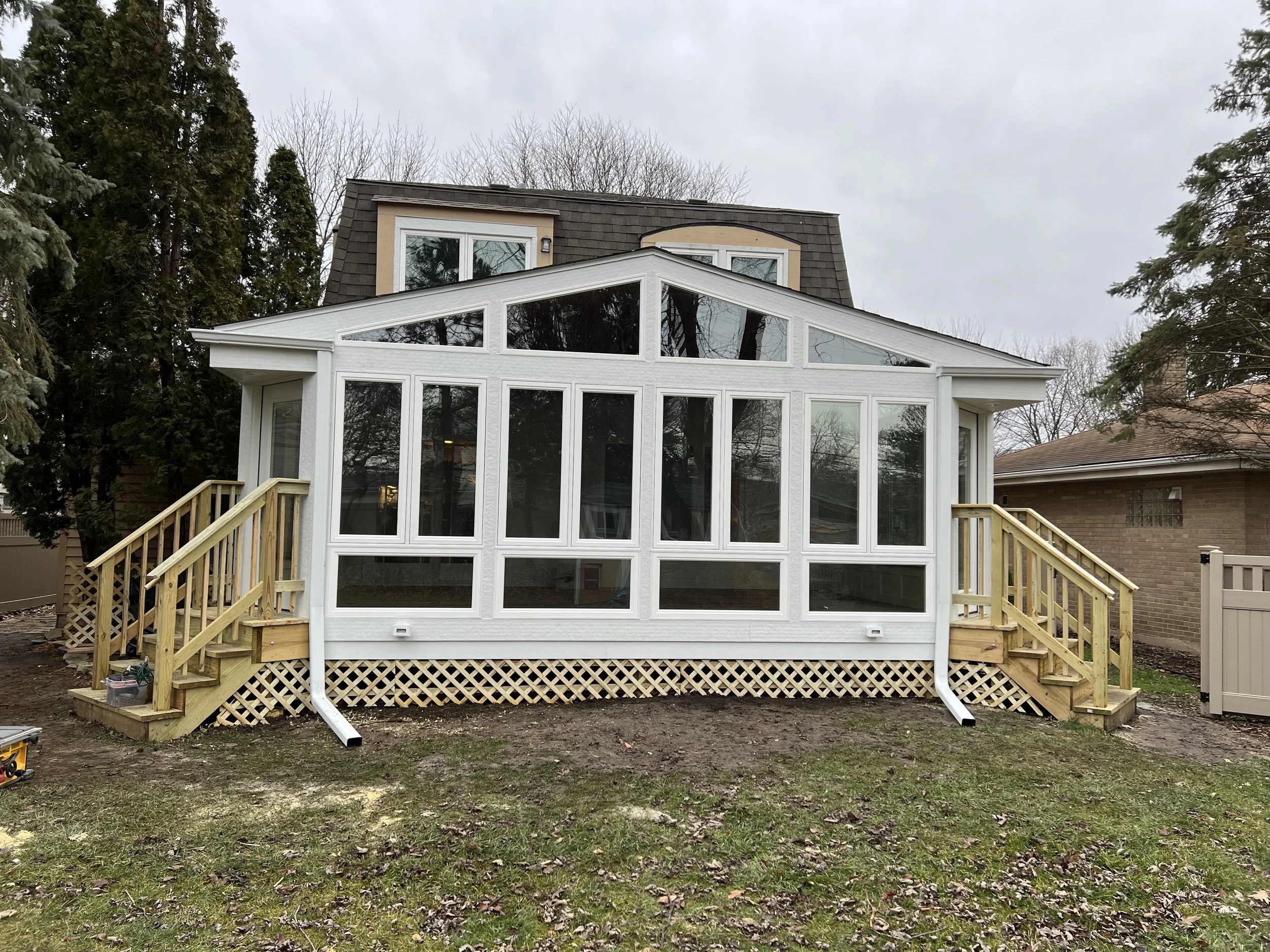 Newly built backyard sunroom with white framing, now under construction, with steps on both sides leading up to it, and a lattice foundation, next to a house with a brown exterior.