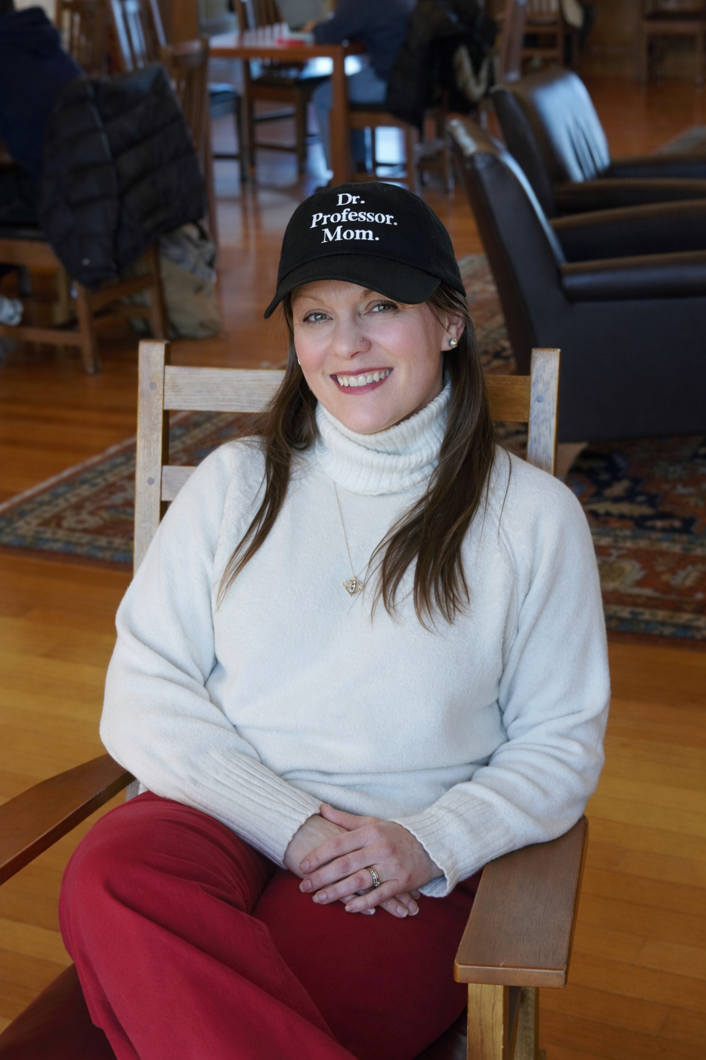 A woman sitting on a wooden chair in a cozy room, wearing a black cap that reads 'Dr. Professor Mom,' a cream-colored turtleneck sweater, and red pants, smiling at the camera.
