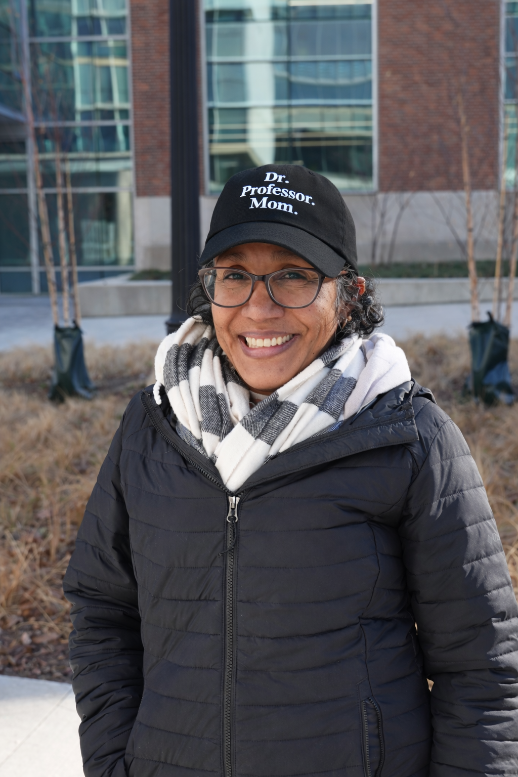 Smiling woman wearing glasses, a black jacket, a black cap with white text, and a white checkered scarf, standing outdoors in front of a modern glass building with some trees and dried grass.