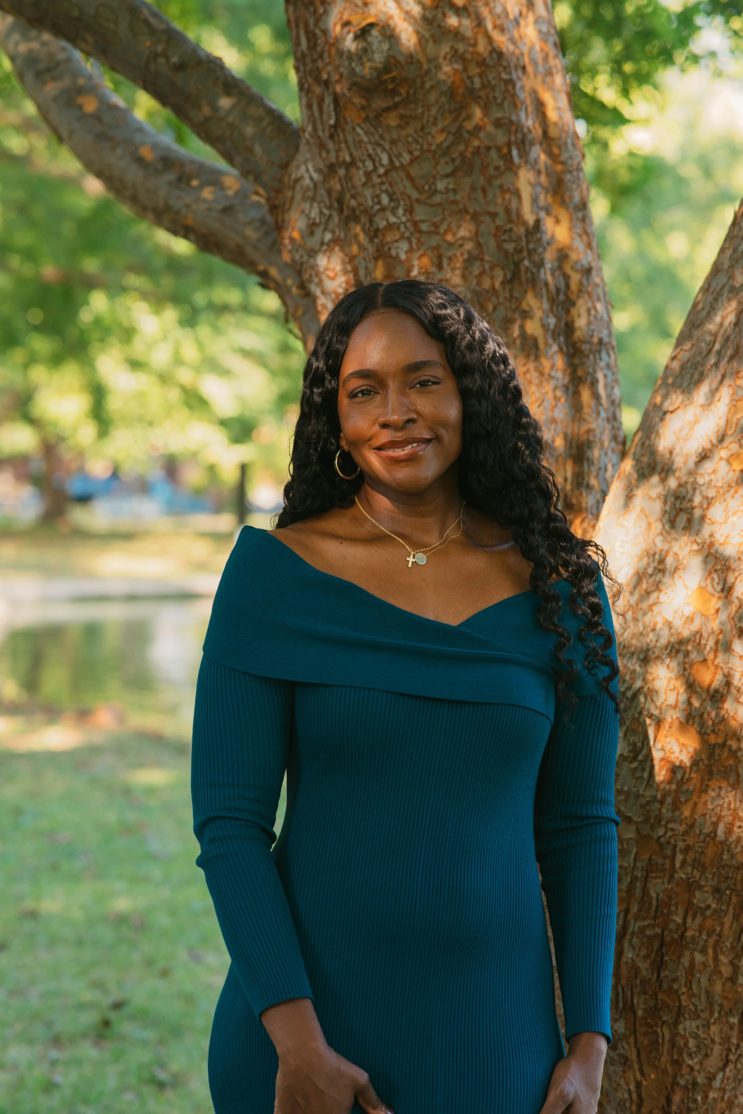 A woman with long, curly black hair stands outdoors next to a large tree, smiling at the camera. She wears a blue off-the-shoulder ribbed dress and gold jewelry, including hoop earrings and layered necklaces. The background features lush green trees and a body of water.