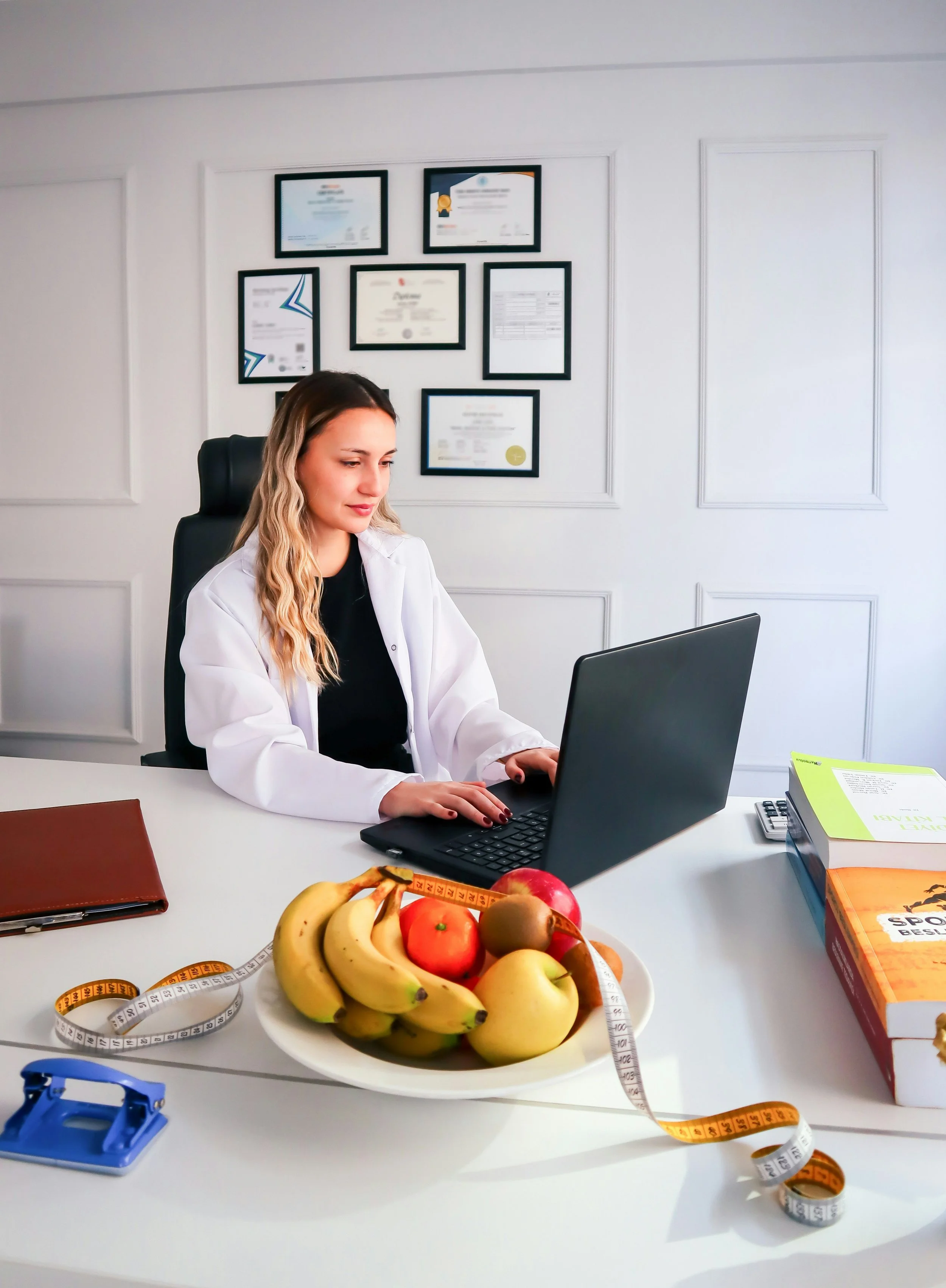 A woman in a white lab coat sitting at a desk with a black laptop, a plate of mixed fruits including bananas and apples, measuring tapes, books, and a calculator in an office with framed certificates on the wall.