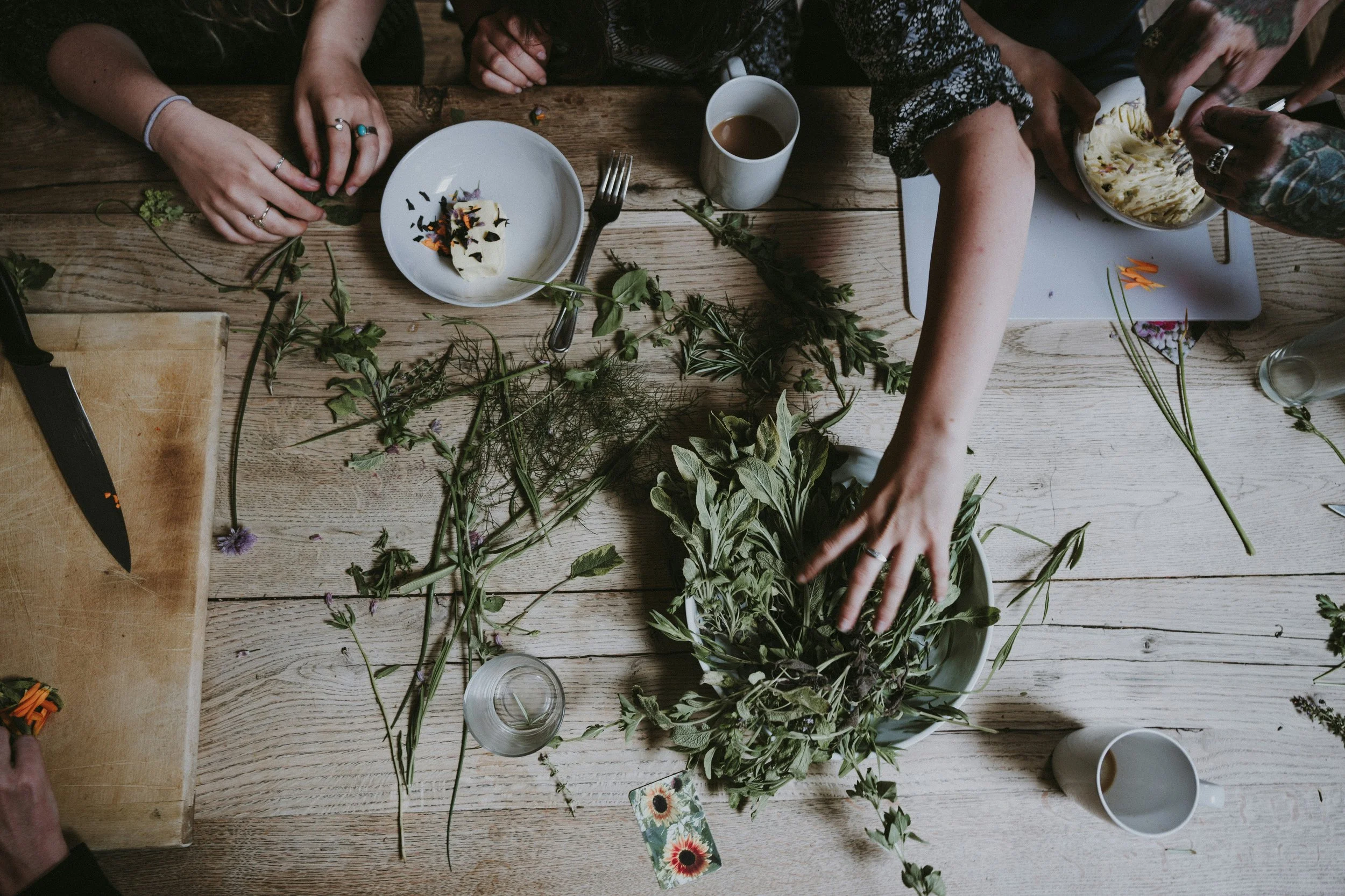 Group of people arranging flowers on a wooden table, with various supplies including a knife, cups, and a knife on a cutting board.