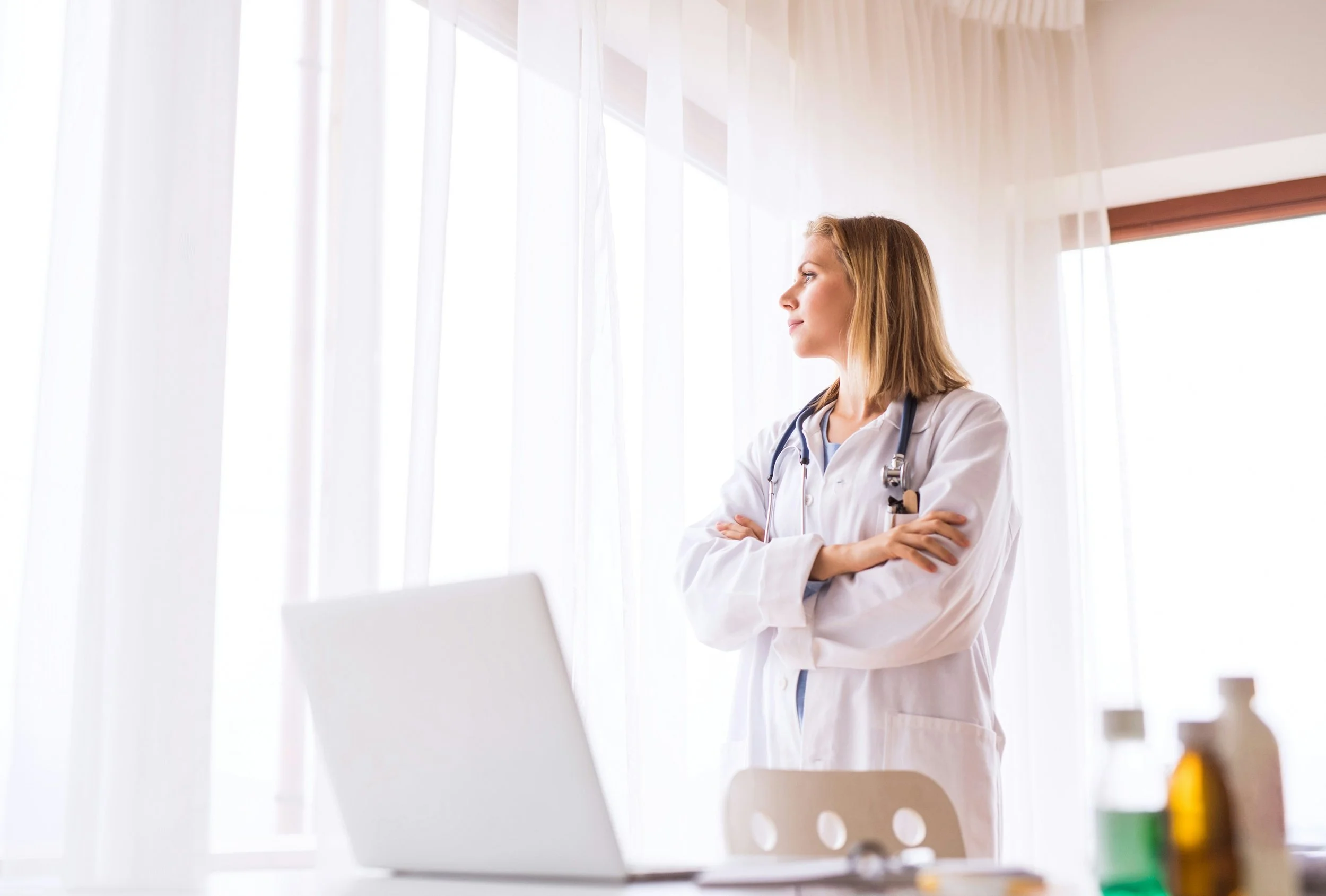 A female healthcare professional, wearing a white coat and stethoscope, standing with arms crossed in a bright room with large windows and sheer curtains.