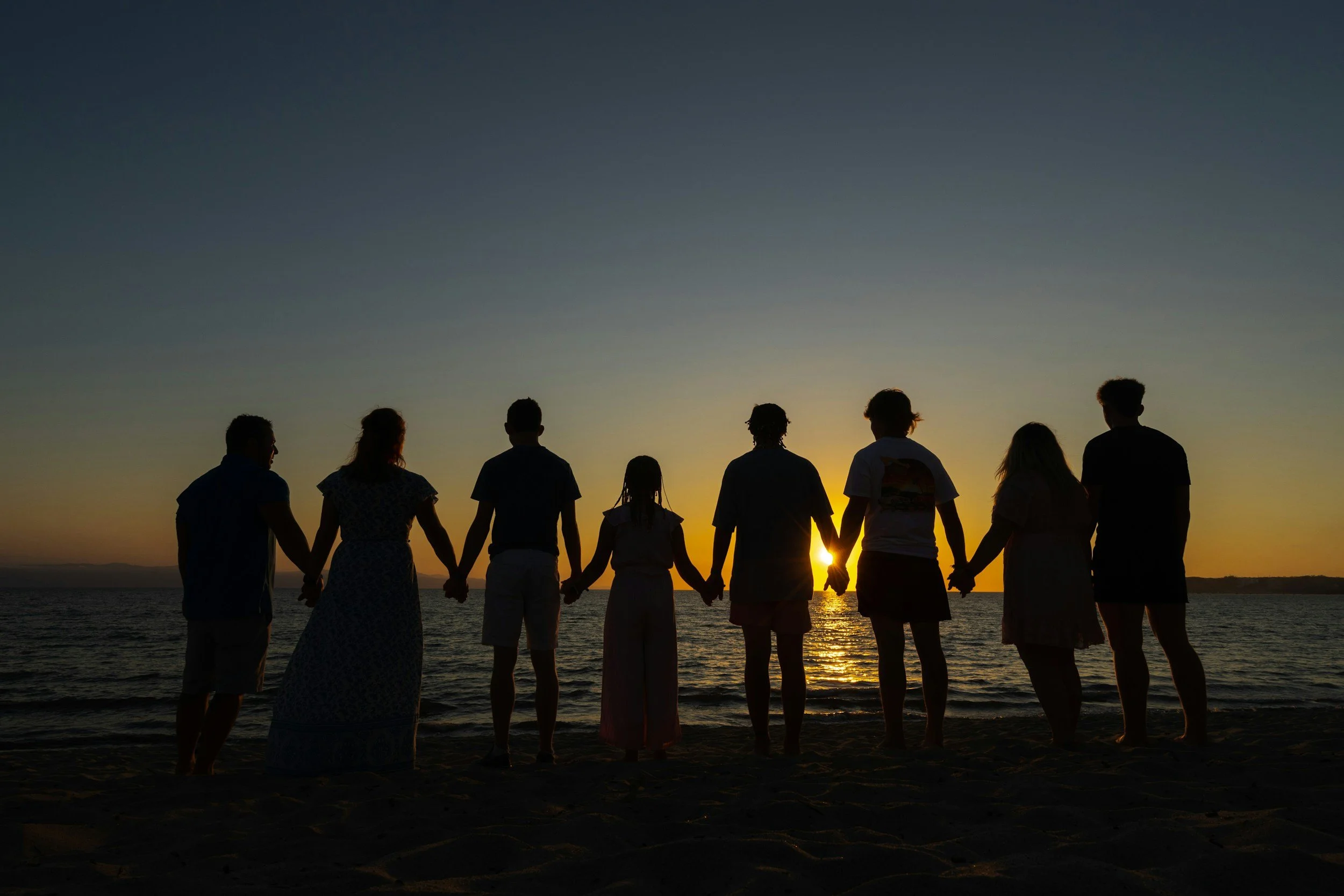 A group of people holding hands on the beach during sunset, facing the water with the sun setting in the background.