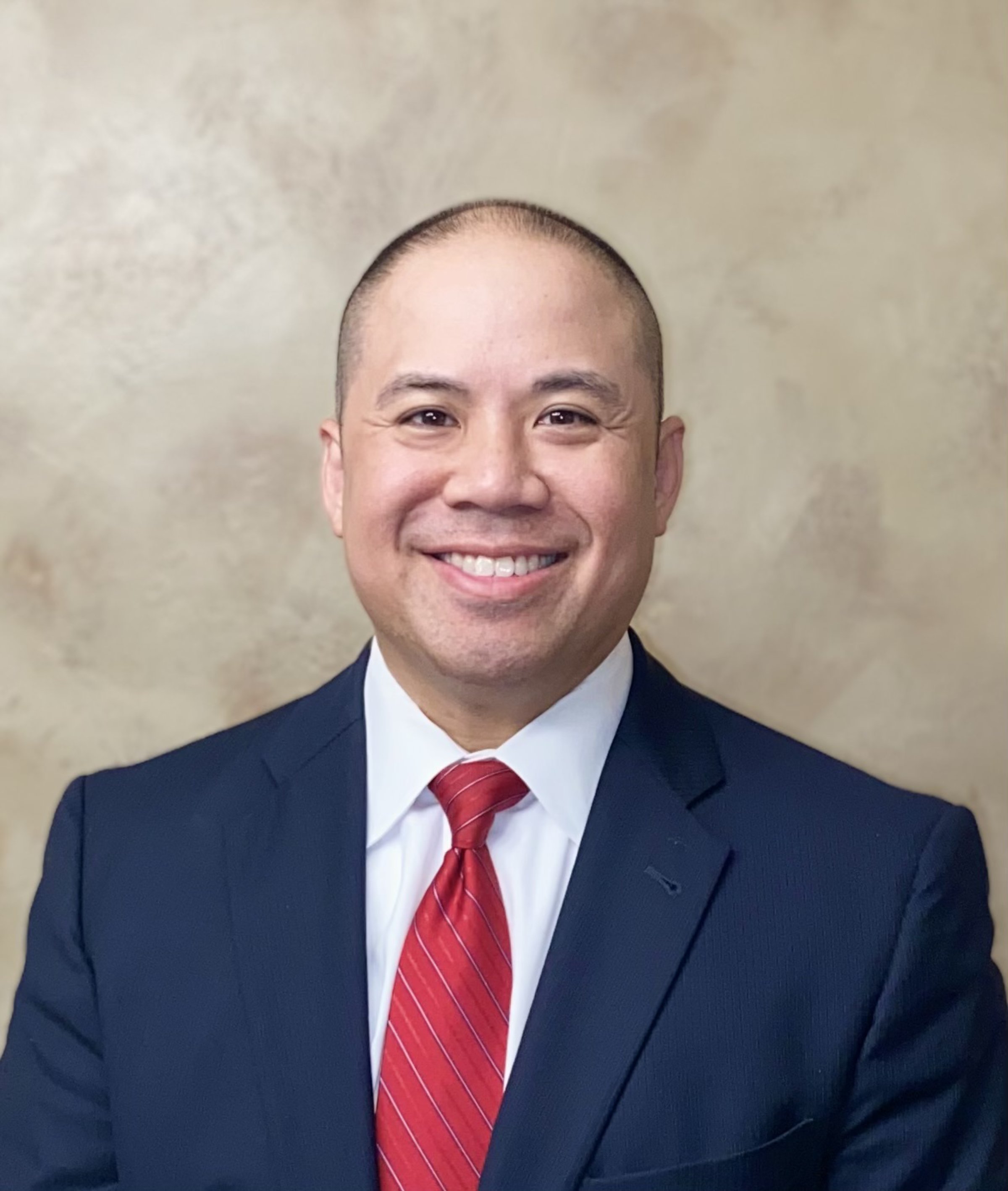 A man in a navy suit, white shirt, and red striped tie smiling against a beige background.