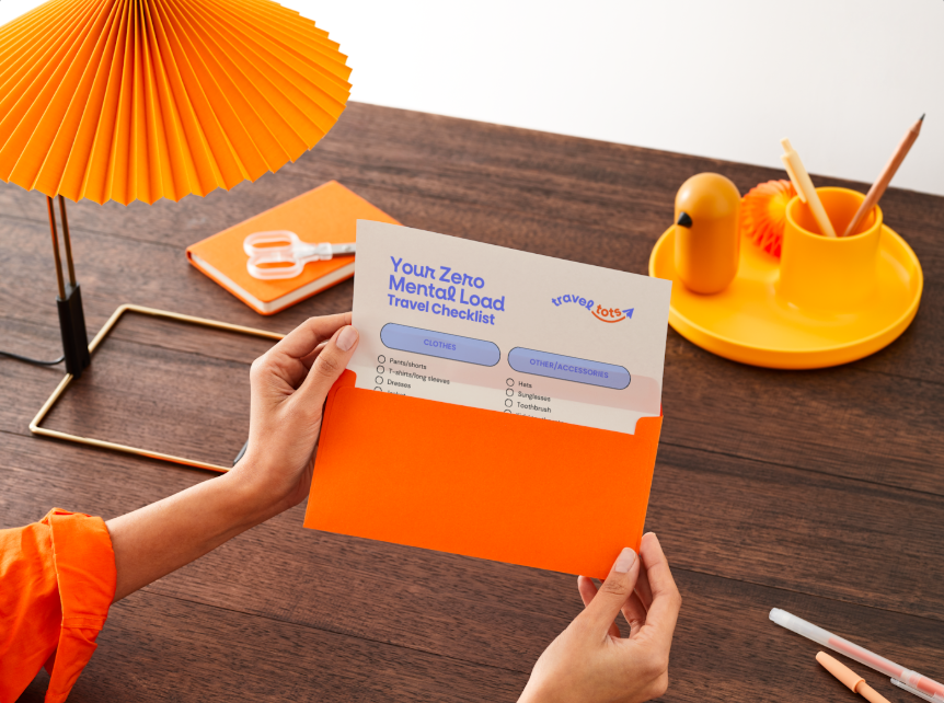 A person holding a travel checklist about mental load with a colorful desk setup including a yellow lamp, a yellow duck décor, and an orange notebook.