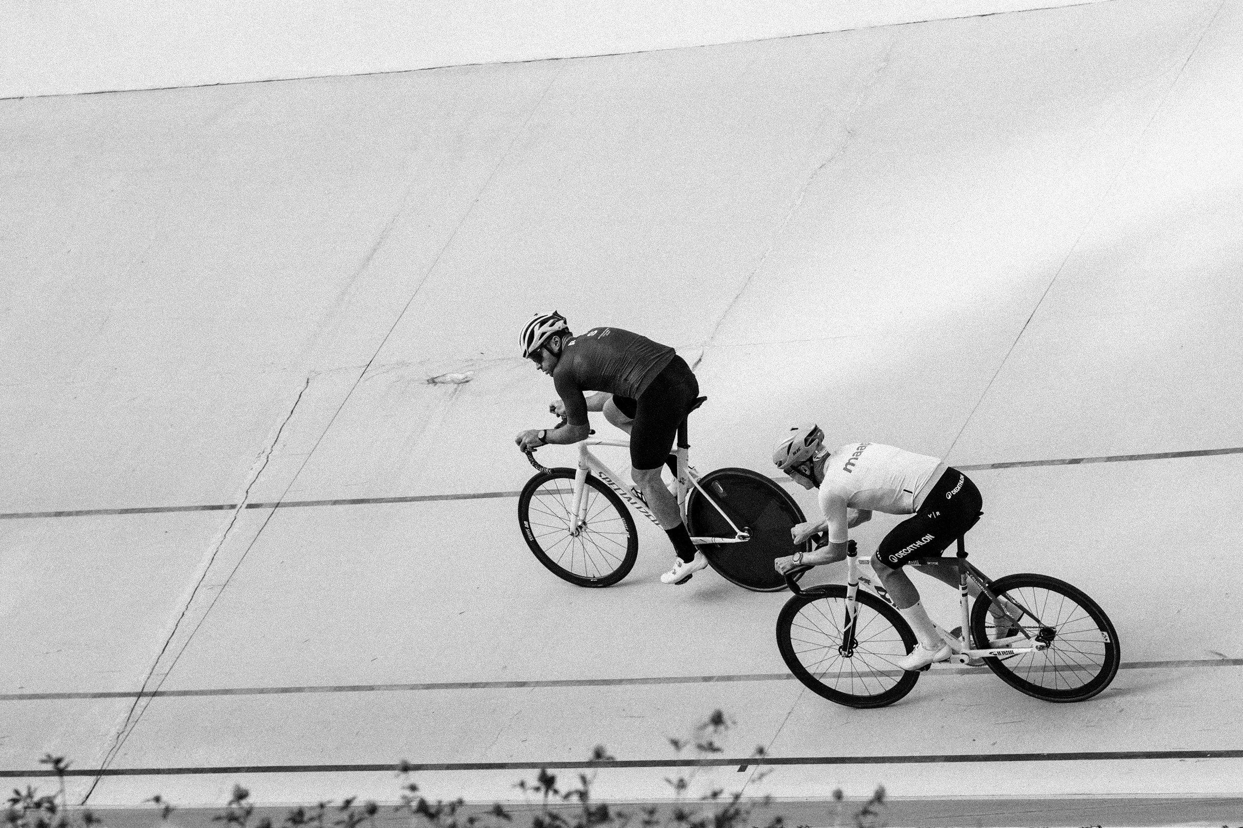 Two cyclists riding on a velodrome track with tilting angles, wearing helmets and sports gear, one cyclist in a white jersey and the other in a dark jersey, with one bike having a disc wheel.