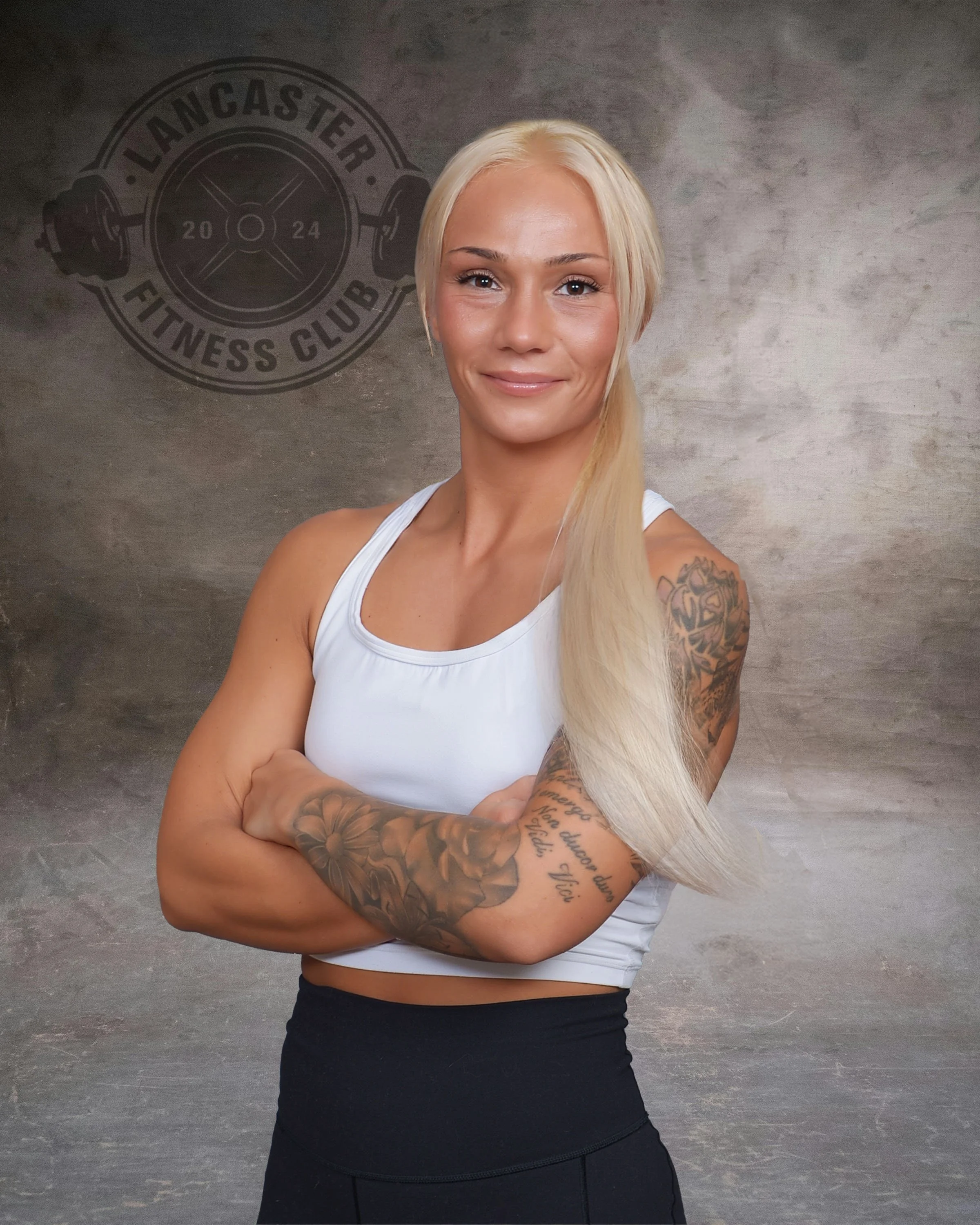 A woman with long blonde hair, tattoos on her arms, wearing a white sleeveless athletic top and black high-waisted workout pants, standing with arms crossed in front of a gray textured wall with a 'Lancaster Fitness Club' logo.