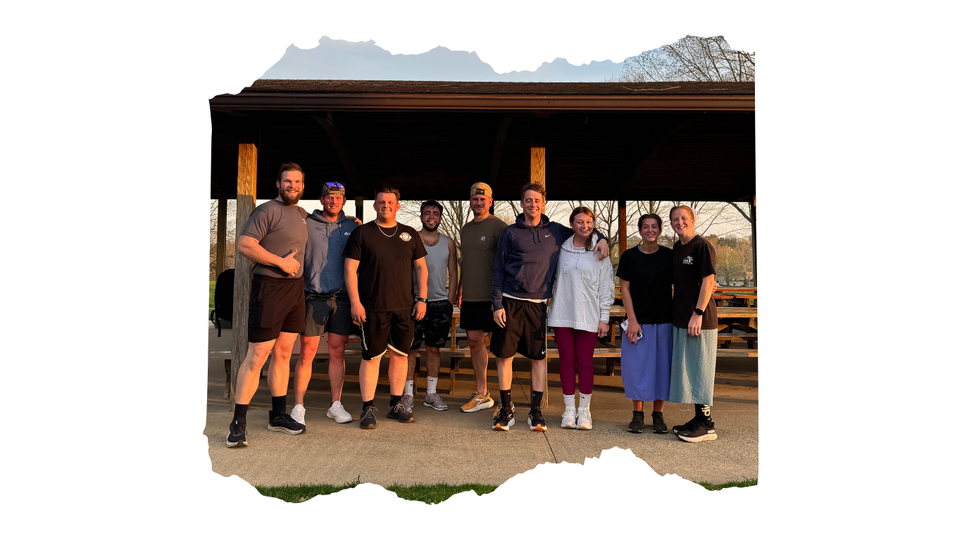 Group of nine people standing outdoors on a paved surface, smiling. They are dressed casually and standing in front of a wooden shelter with picnic tables, with trees and a partly cloudy sky in the background.