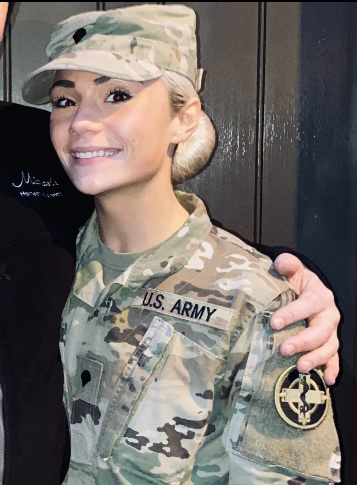 A woman in U.S. Army camouflage uniform smiling, with a cap, standing next to a person with their arm around her shoulder.