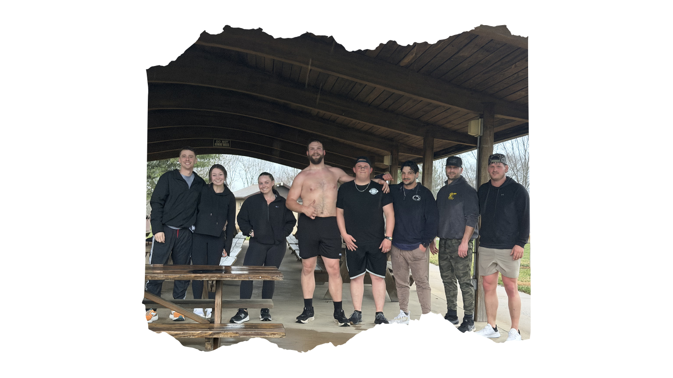 Group of eight young adults standing together outdoors under a wooden shelter, posing for a photo.