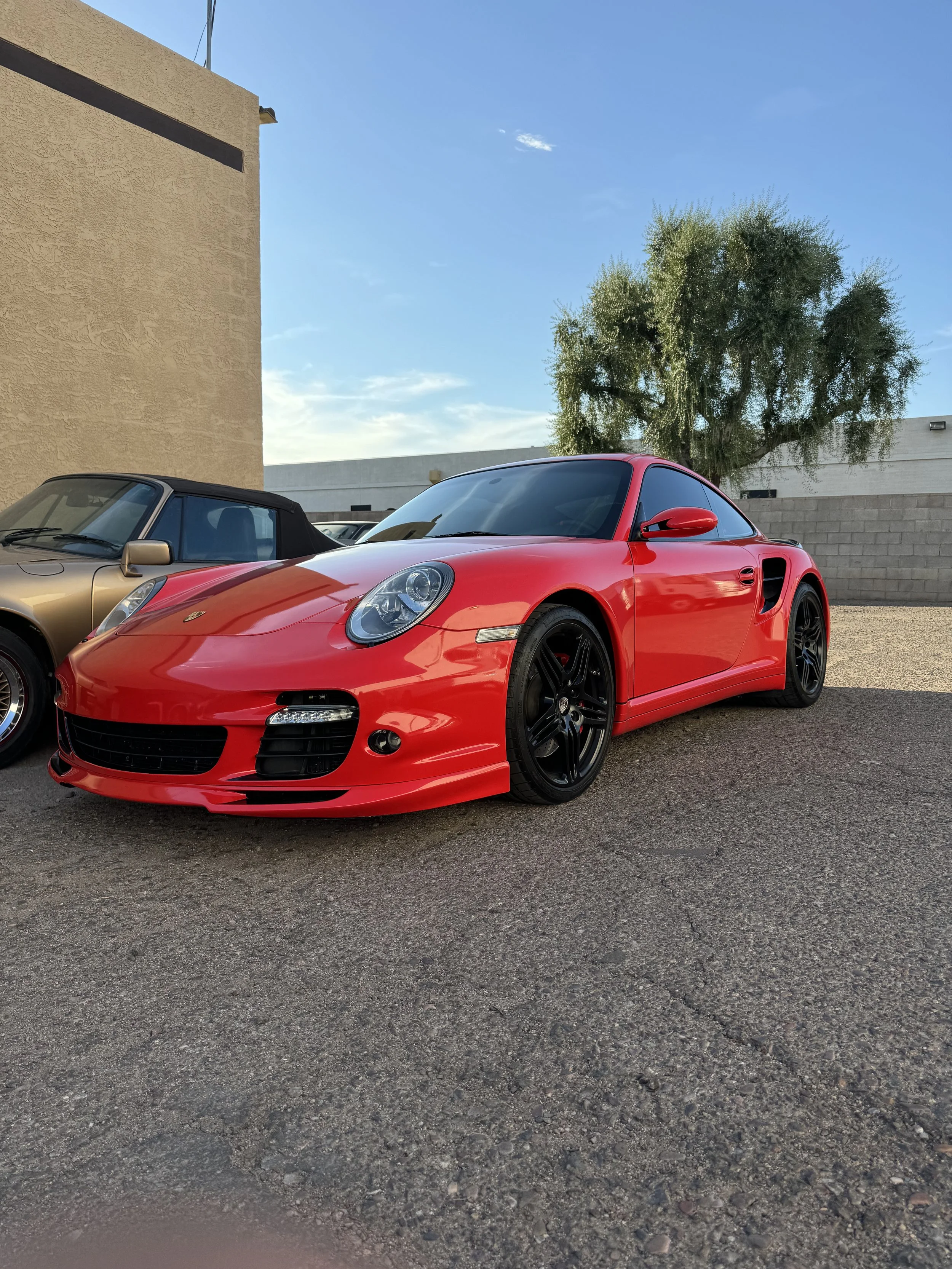 A red Porsche sports car parked on a gravel lot next to a beige classic car, with a tree and building in the background under a blue sky.