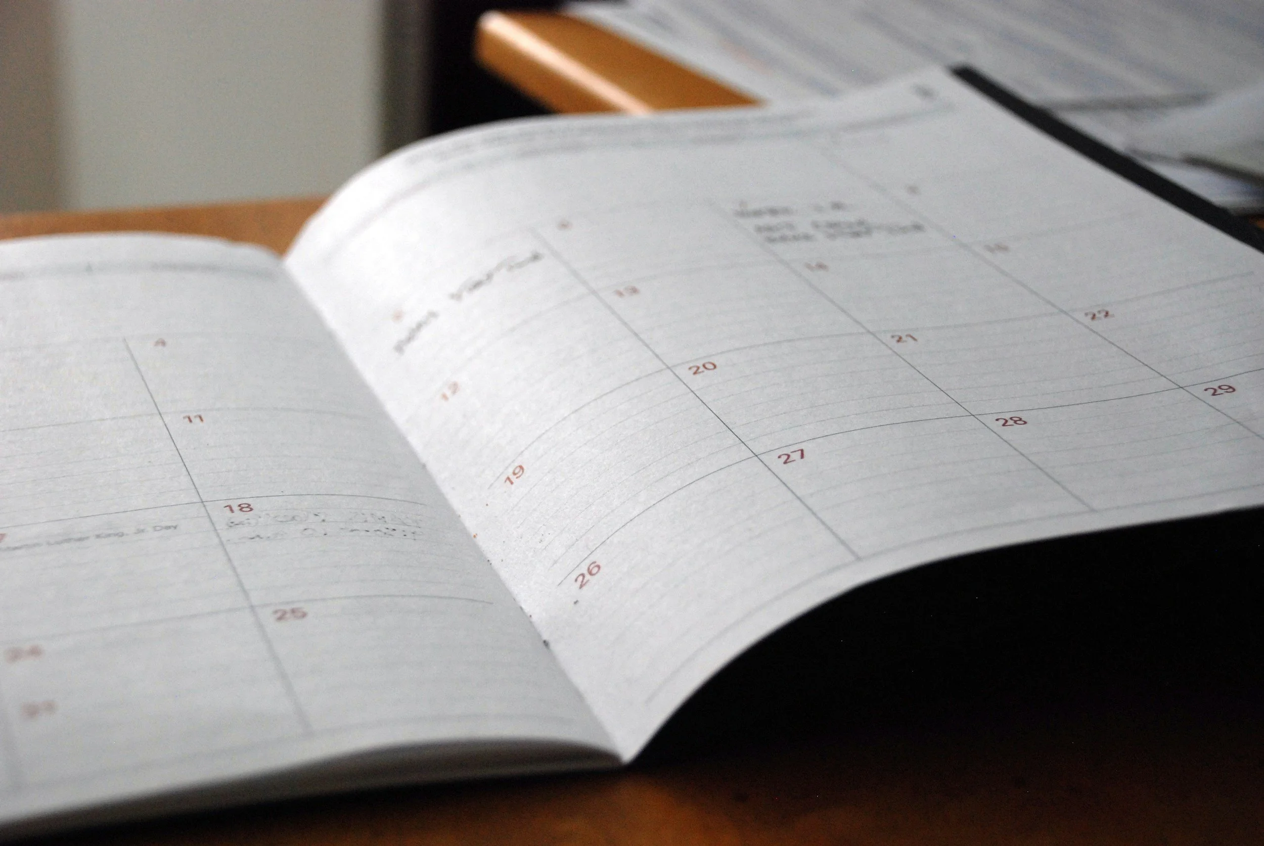 An open planner with a weekly calendar layout on a wooden desk, with additional papers or notebooks in the background.