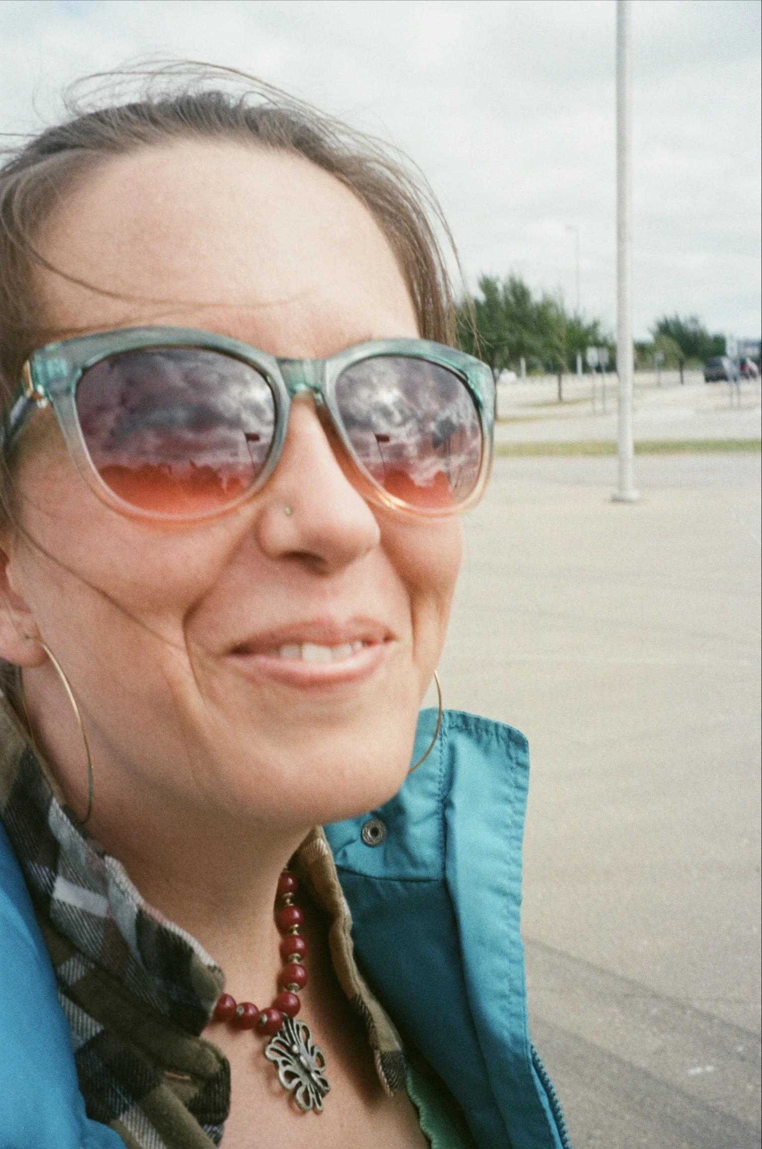 Close-up of a smiling woman wearing large sunglasses, a nose stud, hoop earrings, a necklace with a butterfly pendant, a checkered shirt, and a blue jacket, outdoors on a cloudy day.