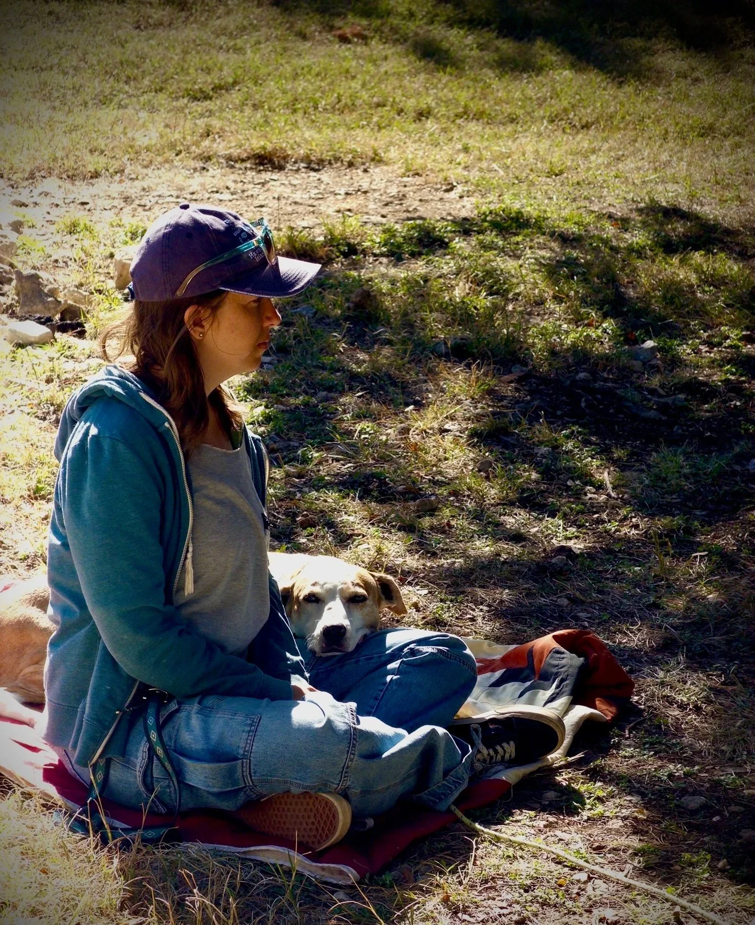 Woman sitting on a blanket with a dog beside her outdoors in a shaded area.