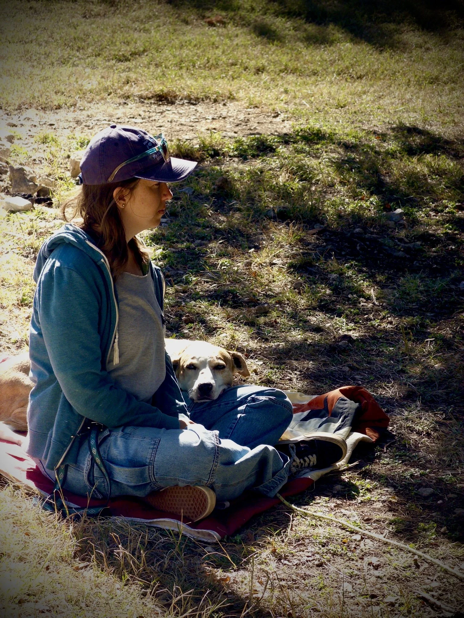 A girl is sitting on a blanket on the ground with her legs crossed and her eyes closed, next to a dog lying down with its head on her lap. The girl is wearing a baseball cap, sunglasses, a hoodie, and jeans. The dog appears relaxed. The scene is outdoors on a grassy area with some shadows and sunlight.