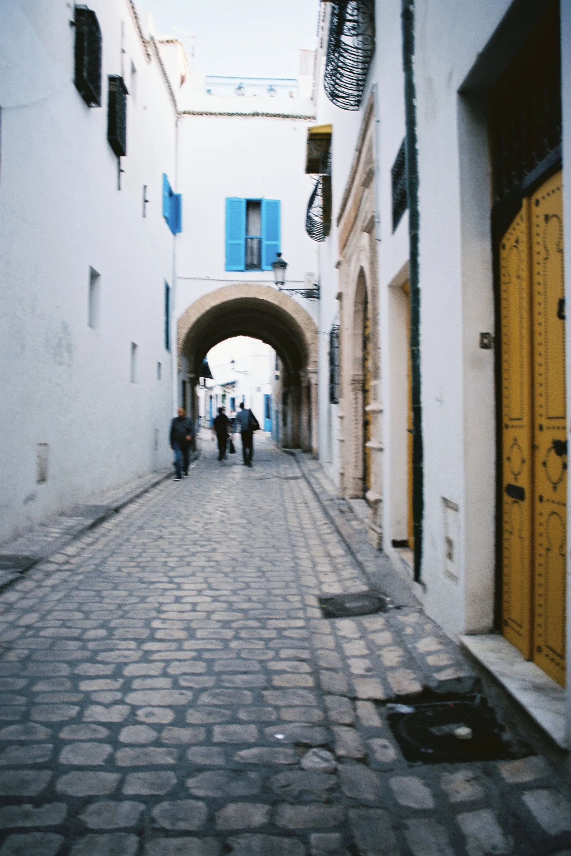 A cobblestone street in a historic European neighborhood, with a stone archway in the distance. White buildings with blue and yellow window shutters. A few people walking.
