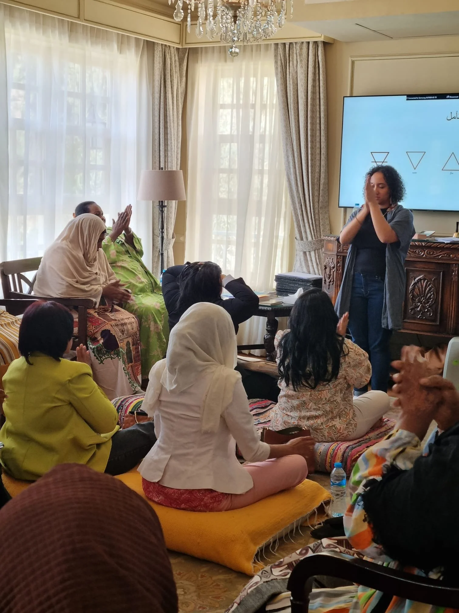 A woman standing in front of a group of seated people during a presentation or workshop in a well-lit room with large windows, curtains, a chandelier, and a chandelier, a large screen displaying diagrams, and various materials and water bottles on a table.