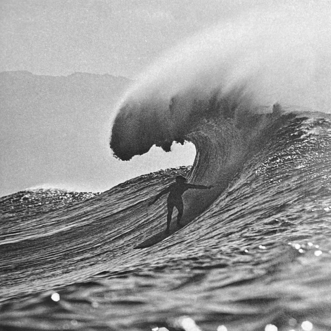 A surfer riding a large wave with the wave curling over, captured in black and white.