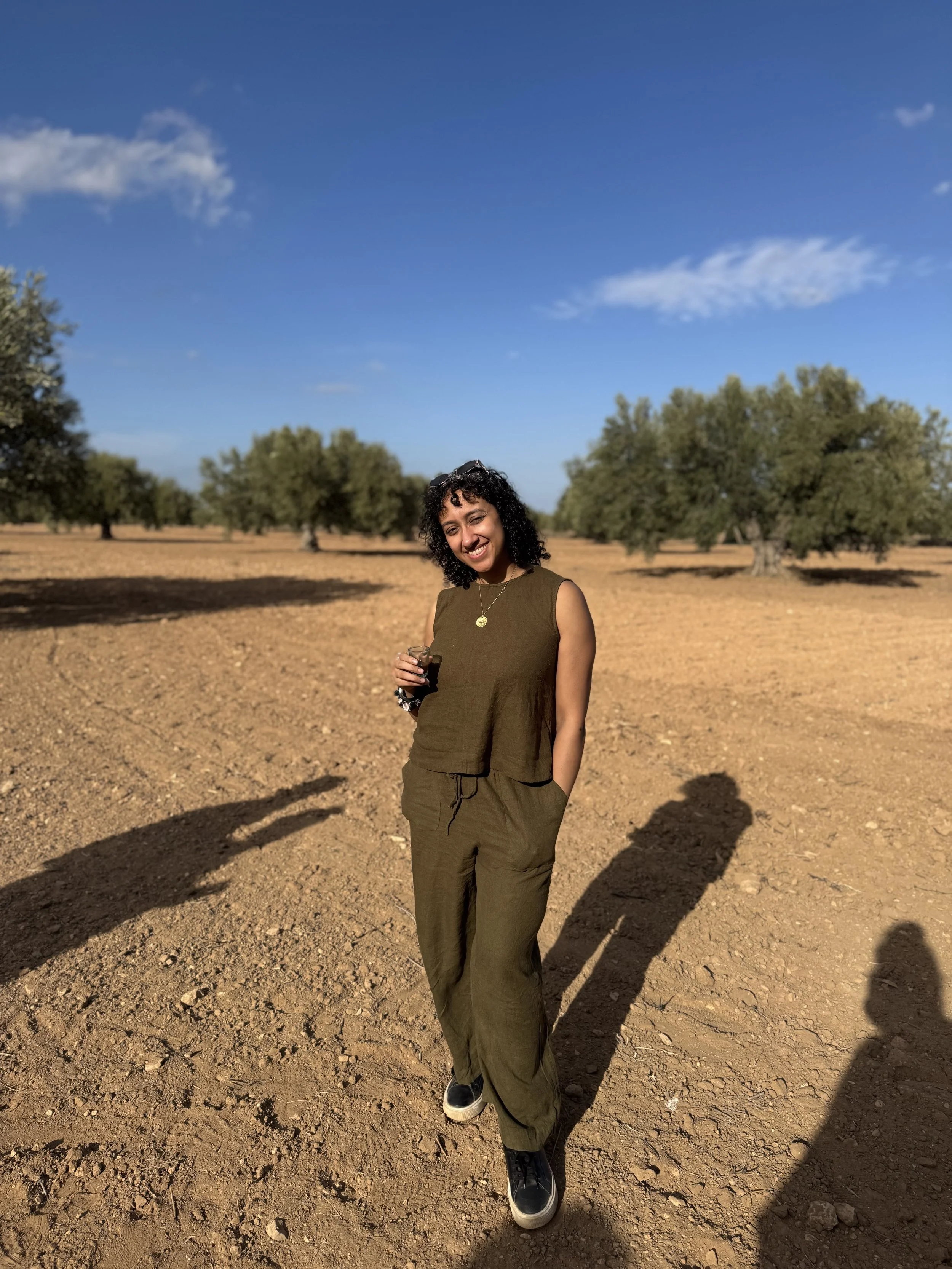 A young woman with curly hair smiling in an olive green sleeveless outfit holding a drink, standing in a dry, open field with scattered trees and a bright blue sky.
