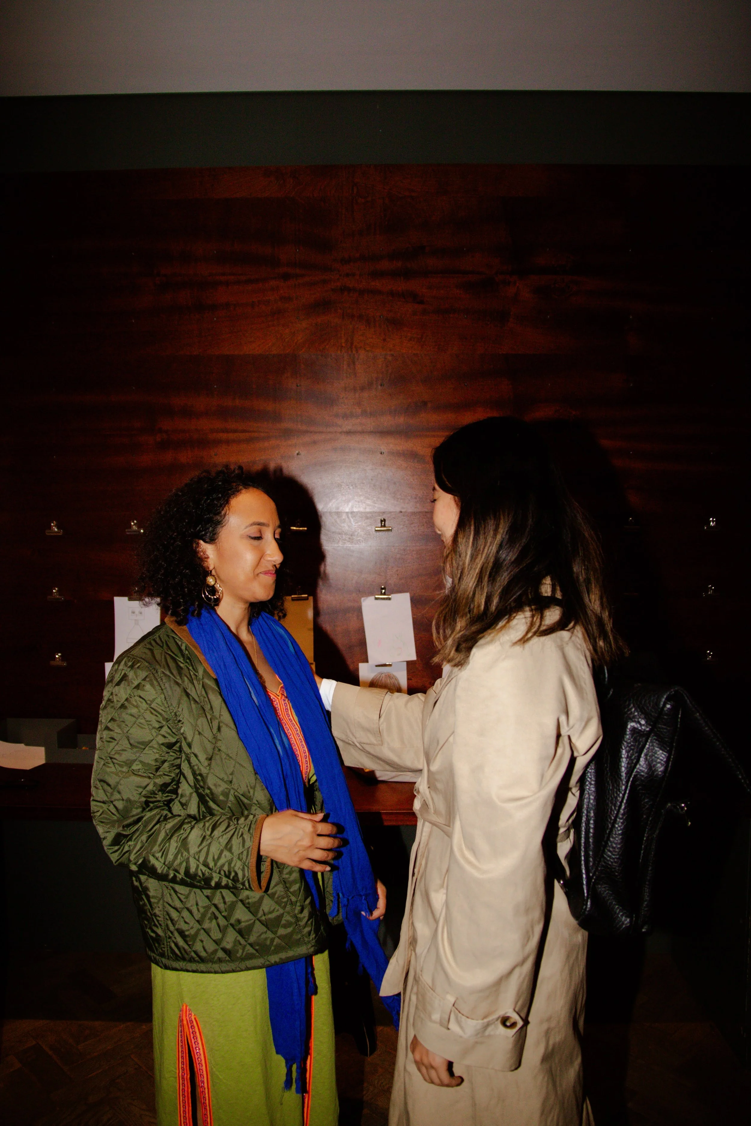 Two women engaged in a warm hug in an indoor space with a dark wooden wall and papers attached to it, one is wearing a green quilted jacket and a vibrant green skirt with red and orange accents, the other is in a beige trench coat with a black backpack.
