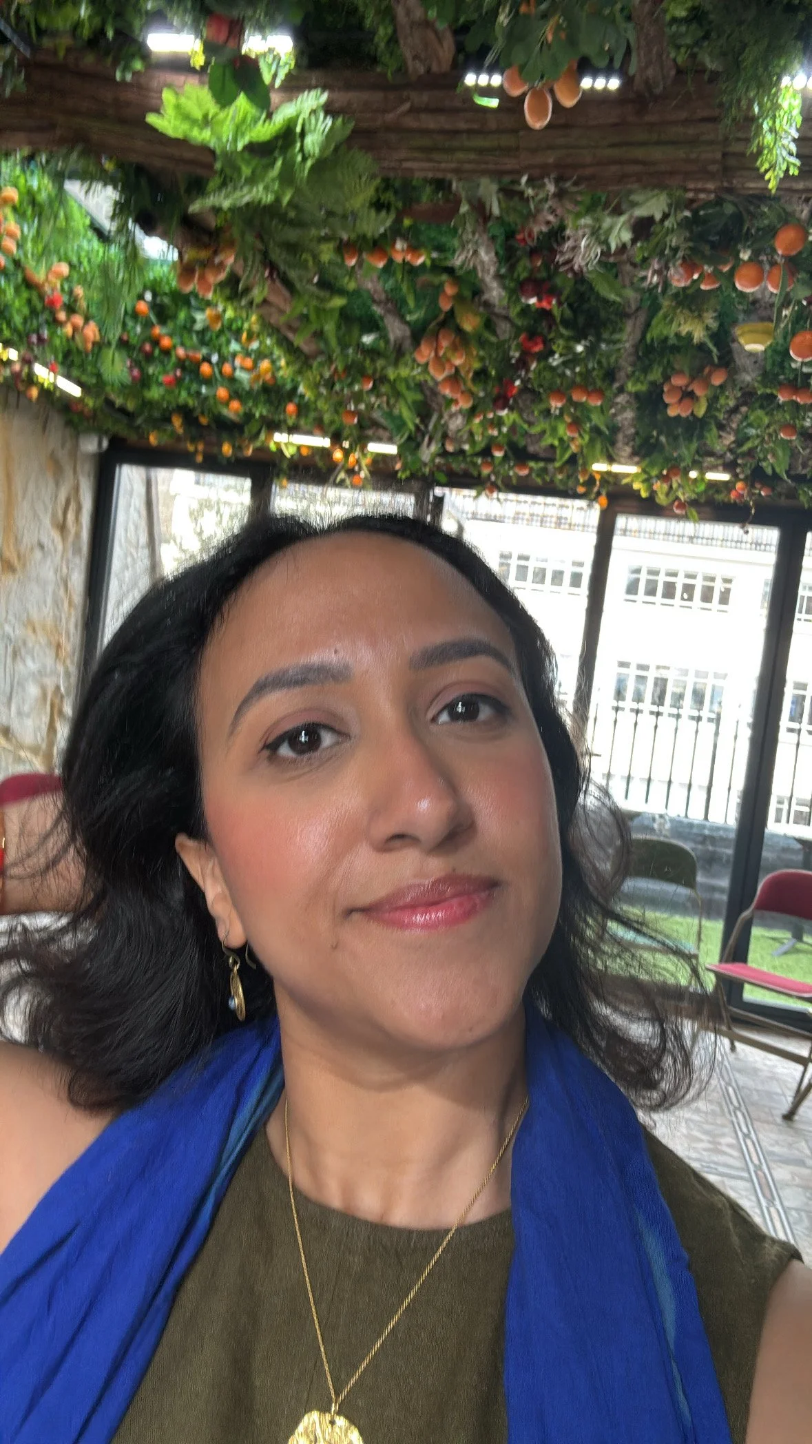 A woman with dark hair, wearing a gold necklace and earrings, smiling in a room with large windows. Overhead, there is a decorative ceiling with artificial green leaves, orange berries, and hanging orange fruits.