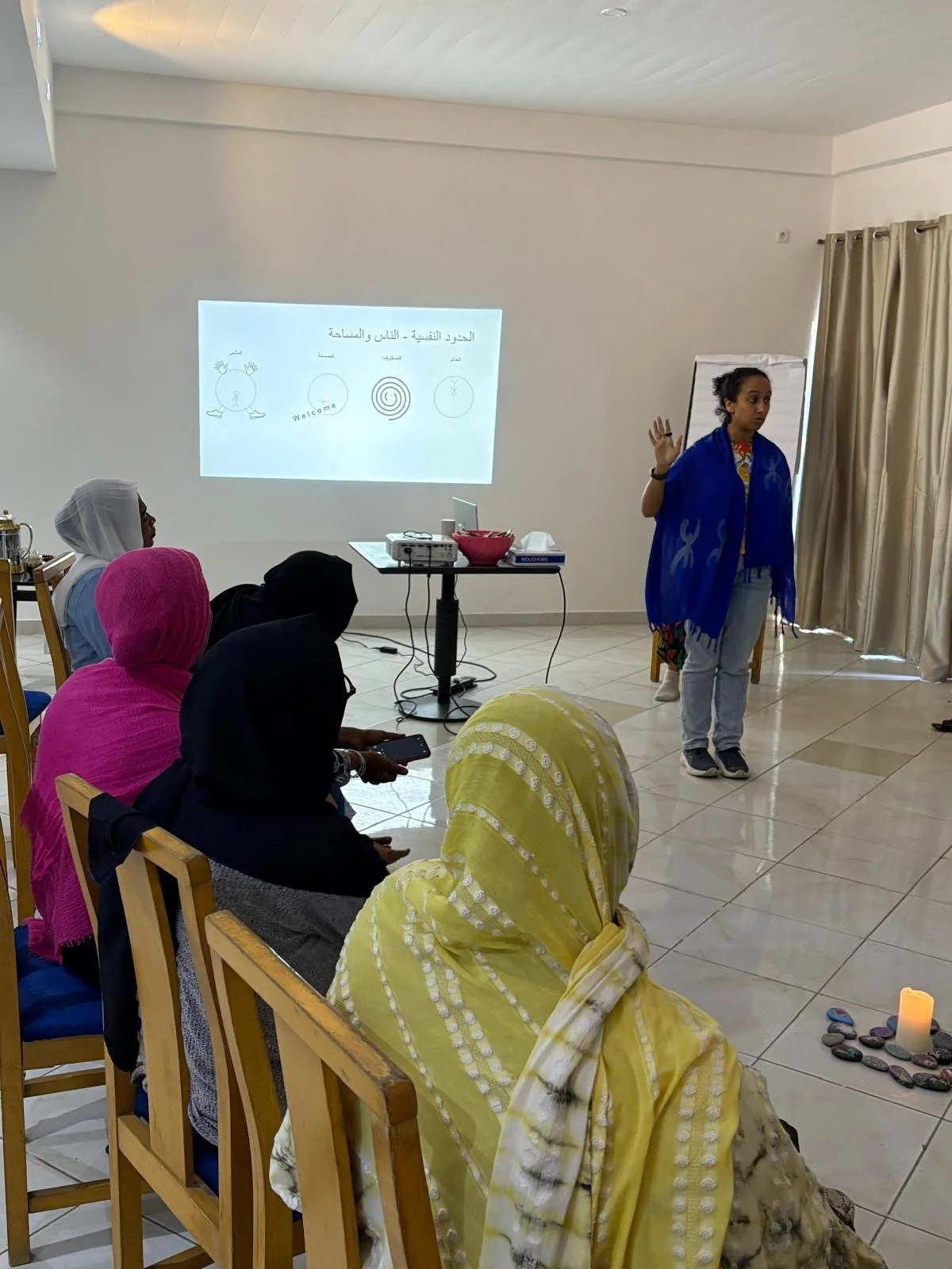 A woman is giving a presentation to a group of seated women inside a room, with a projection screen displaying Arabic text and graphics. The women are wearing colorful headscarves, and the presenter is wearing a blue scarf.