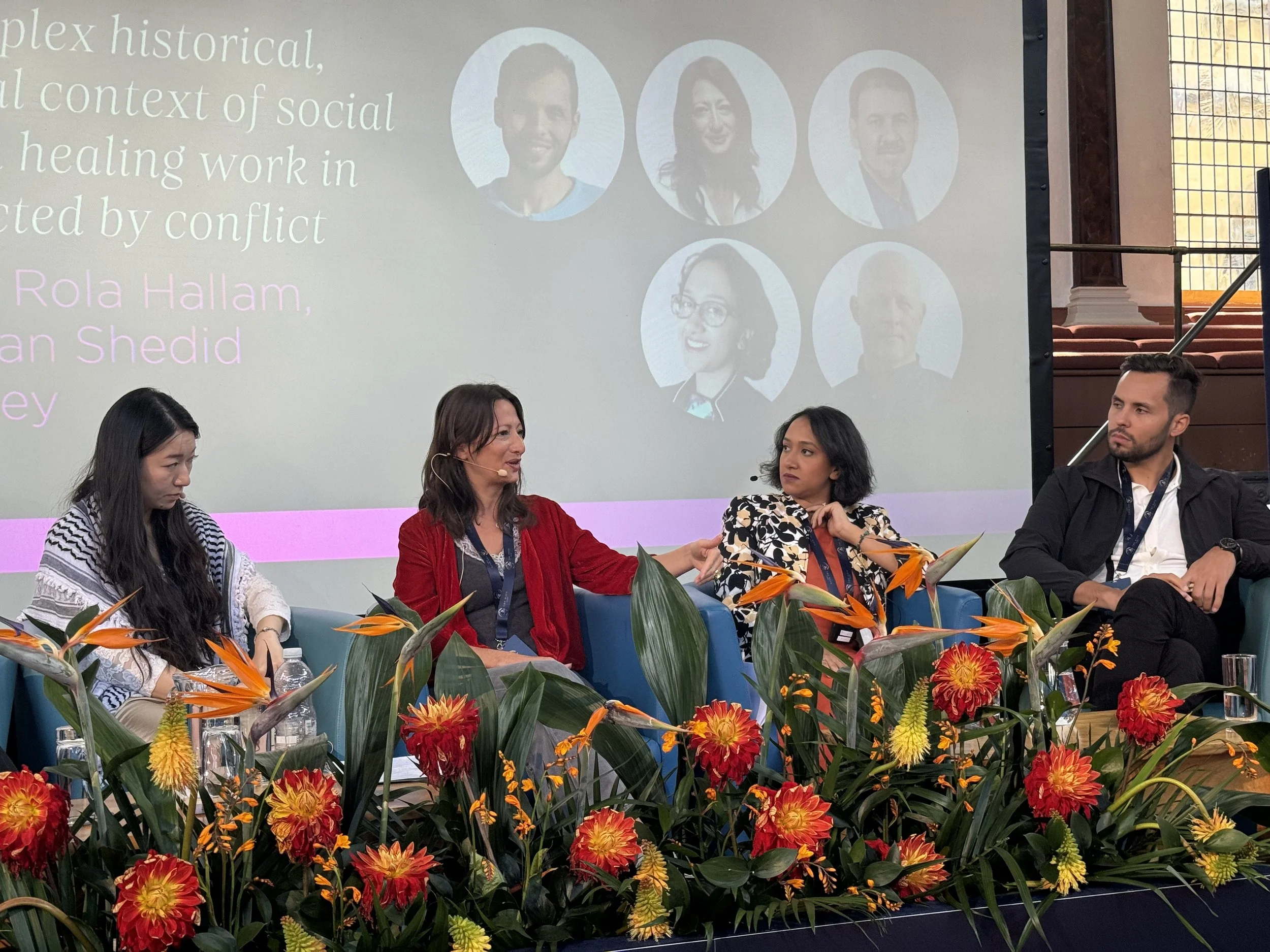A panel discussion with four diverse people seated on a stage decorated with orange and red flowers. Behind them is a large screen displaying circular headshots of five more speakers and some partially visible text.