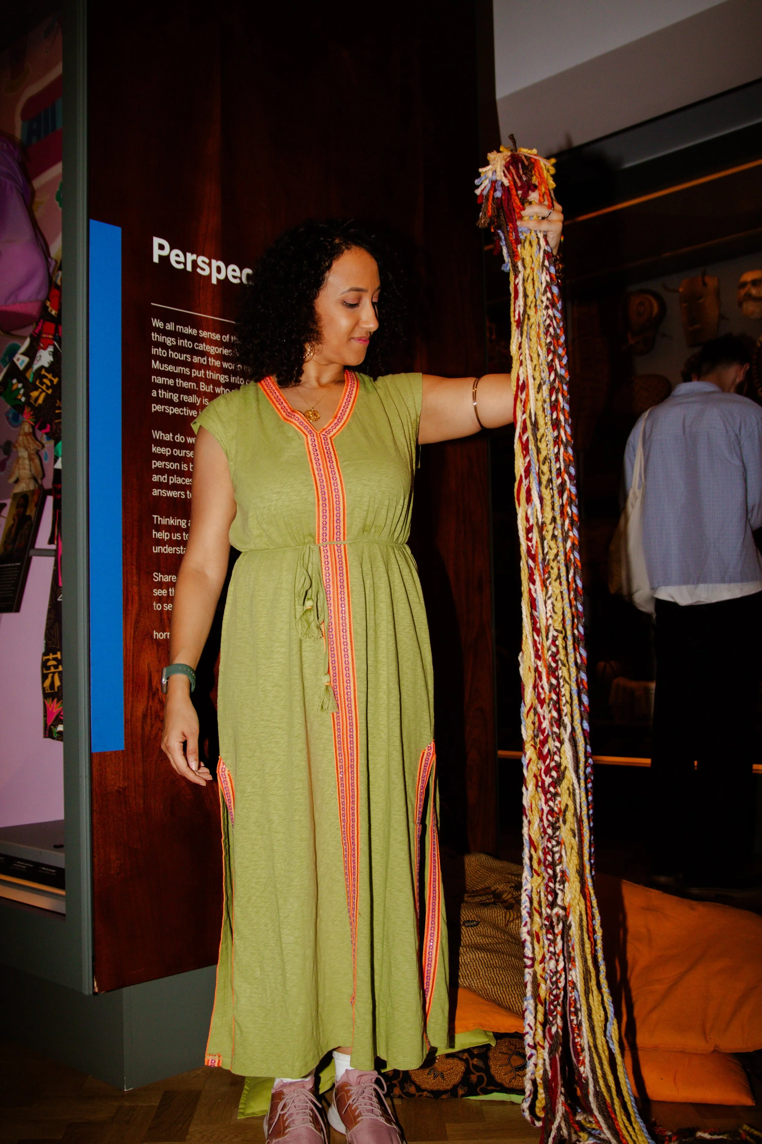 Woman in green dress holding a long, colorful woven textile at a museum or gallery exhibit.