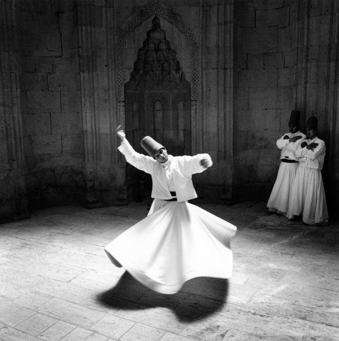 A Sufi whirling dervish performing a traditional dance in a stone-walled room, with two dervishes standing in the background.