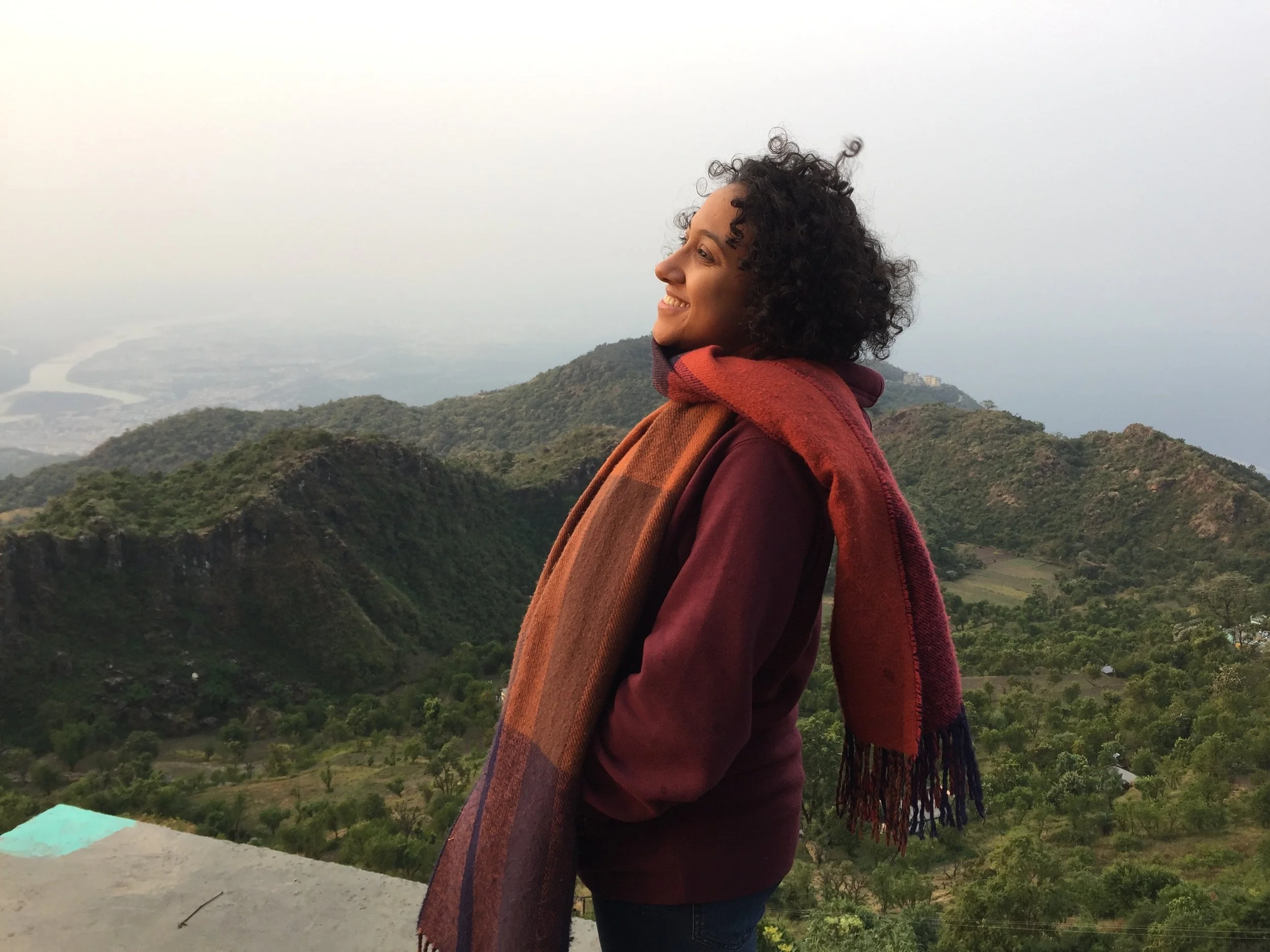 A woman standing on a hilltop with mountains and a river in the background, smiling and looking to the side.