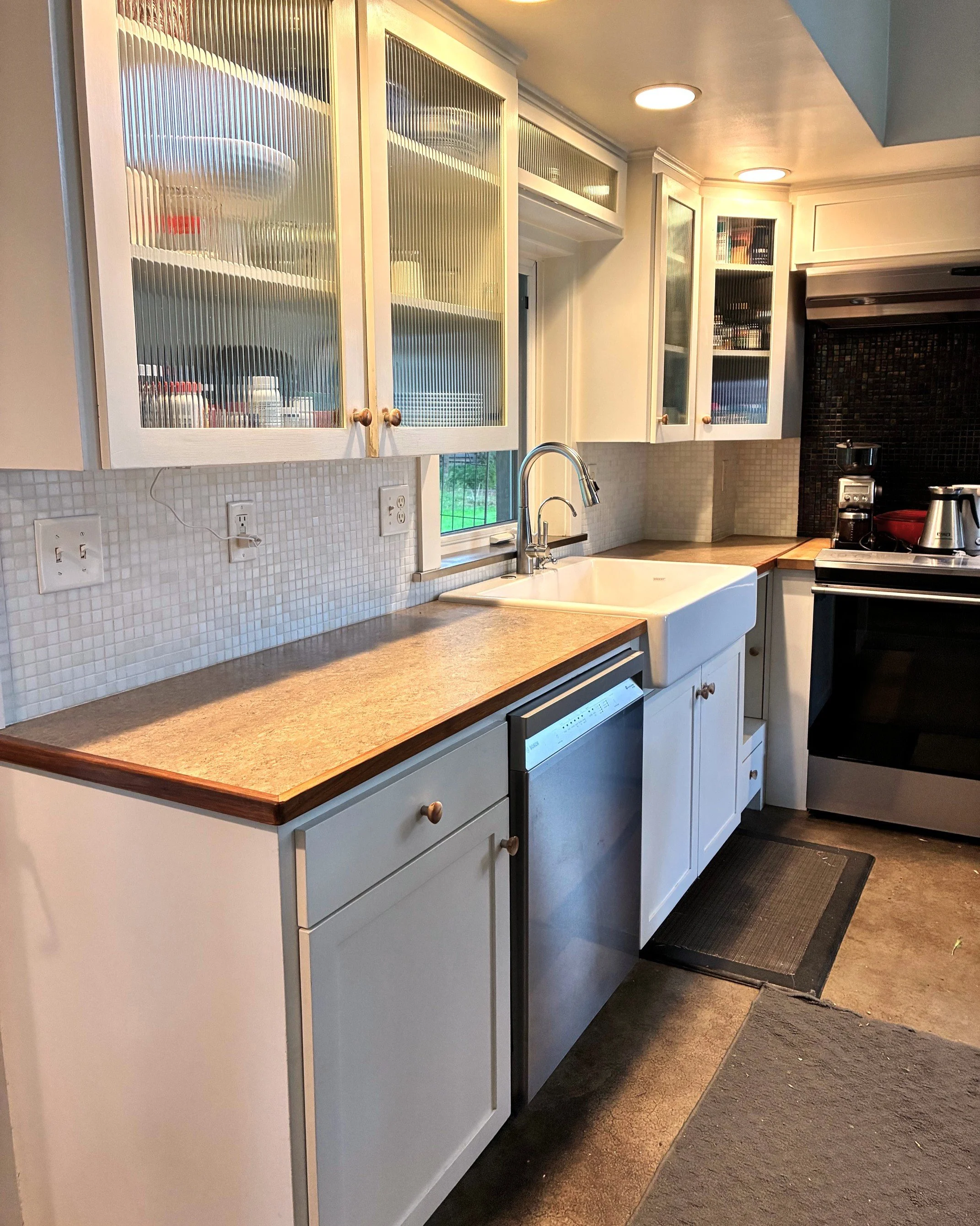 Kitchen with white cabinets, a beige countertop, a white farmhouse sink, a dishwasher, a window above the sink, and black appliances including a stove. The cabinets have glass doors with visible dishes inside.