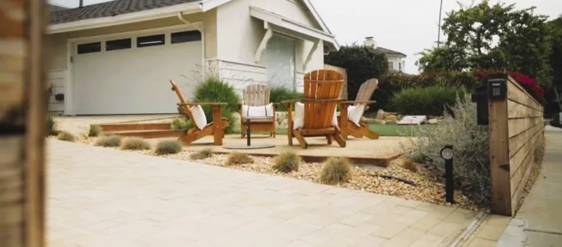 Front yard of a house with a patio area featuring four Adirondack chairs and two wooden chairs, surrounded by gravel and desert plants.
