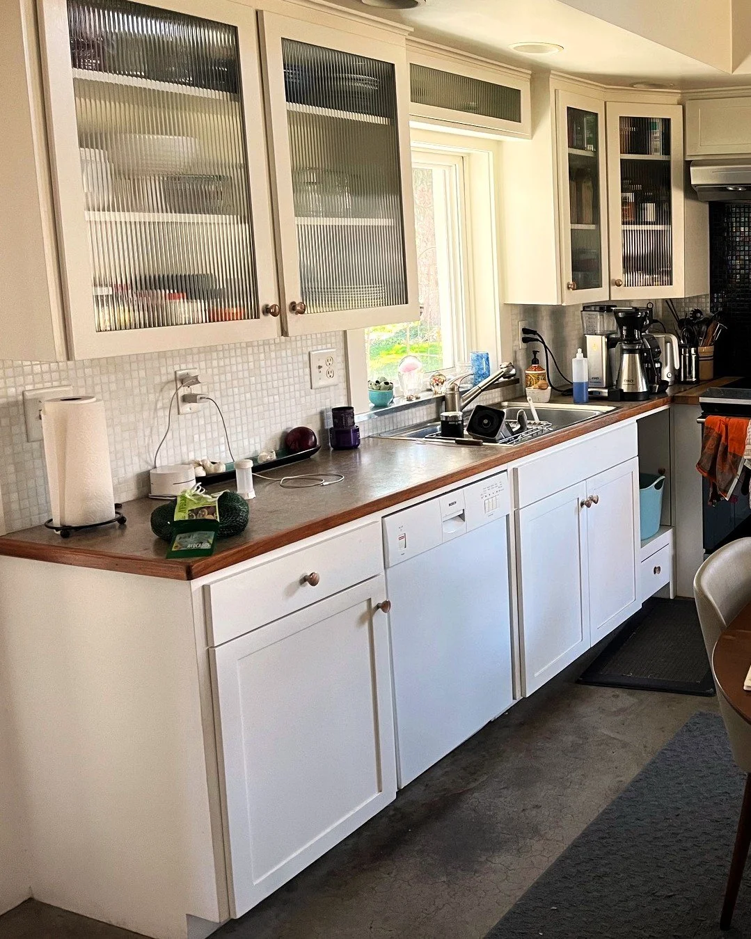 Kitchen with white cabinets, a window above the sink, various small appliances on the countertop, and kitchenware inside glass-front cabinets.
