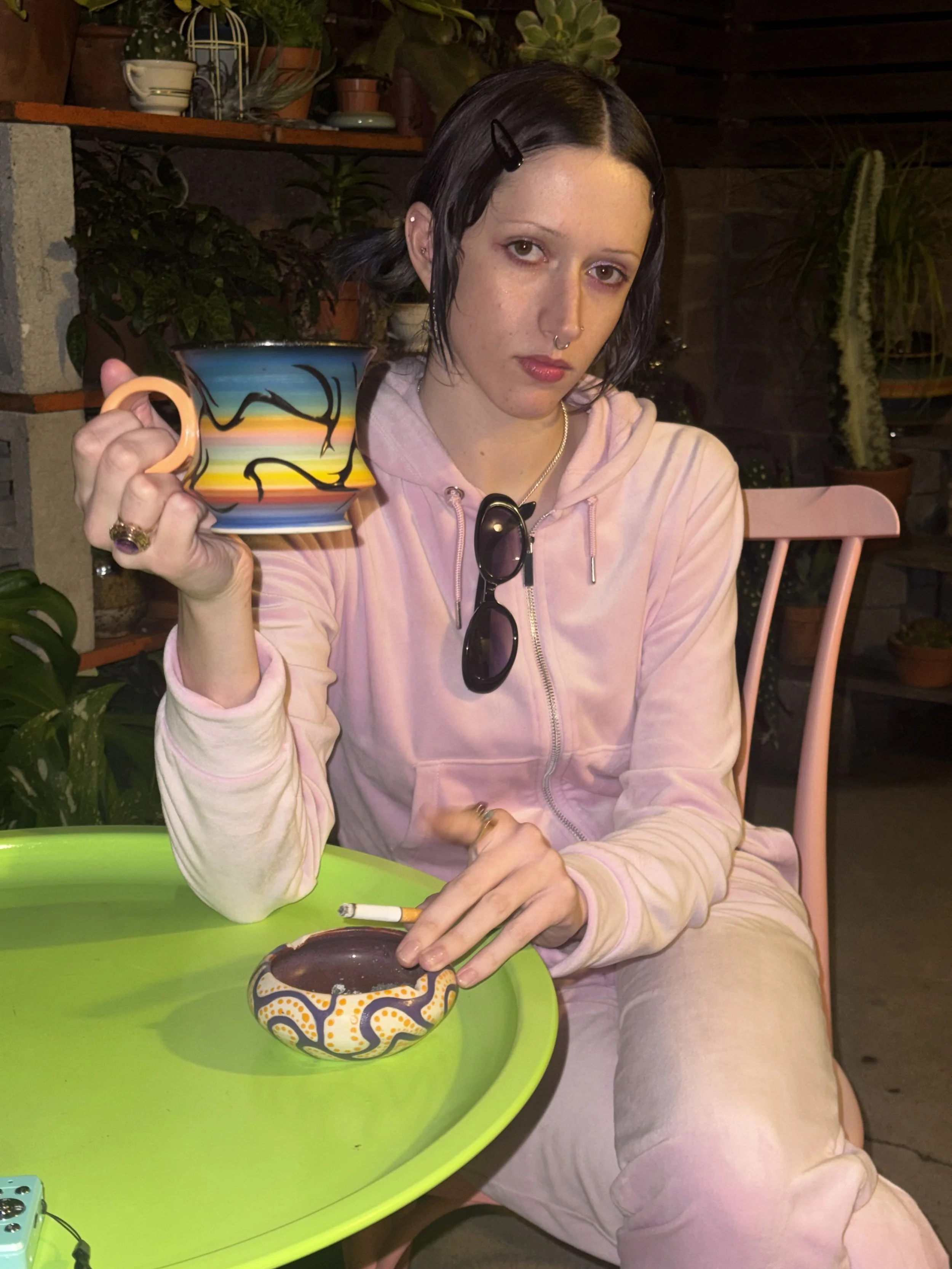 A young woman with short black hair, wearing a pink hoodie and beige pants, sitting at a pink chair. She is holding a colorful mug with a sunset and mountain design in her right hand, with sunglasses hanging from her hoodie. There is a small decorative bowl with a pattern on a bright green table, with a cigarette resting on the bowl. The background includes various potted plants and wooden shelving.