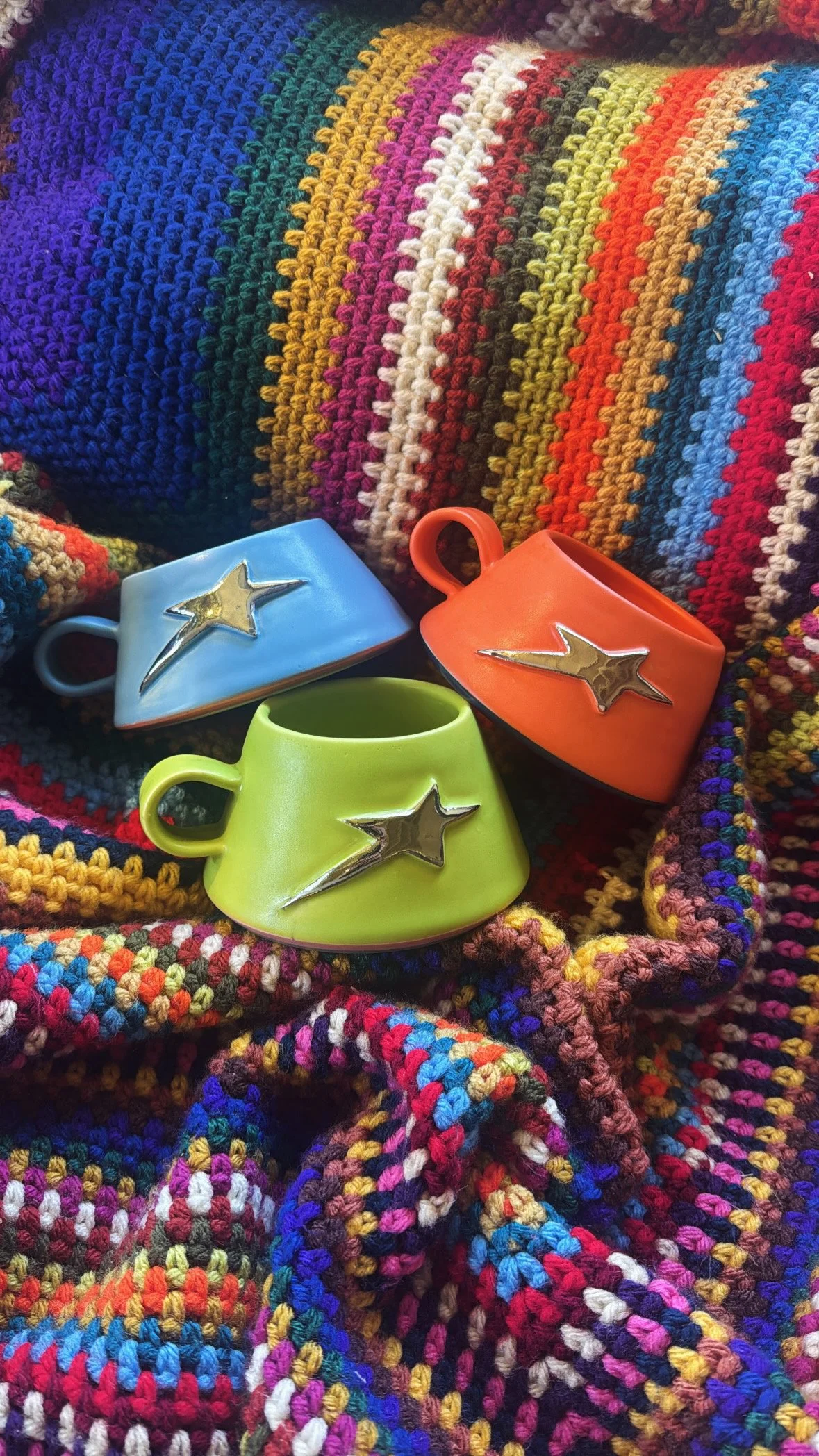 Three colorful ceramic teacups, one blue, one orange, and one lime green, each with a silver star and shooting star design, are placed on a multicolored crocheted blanket.