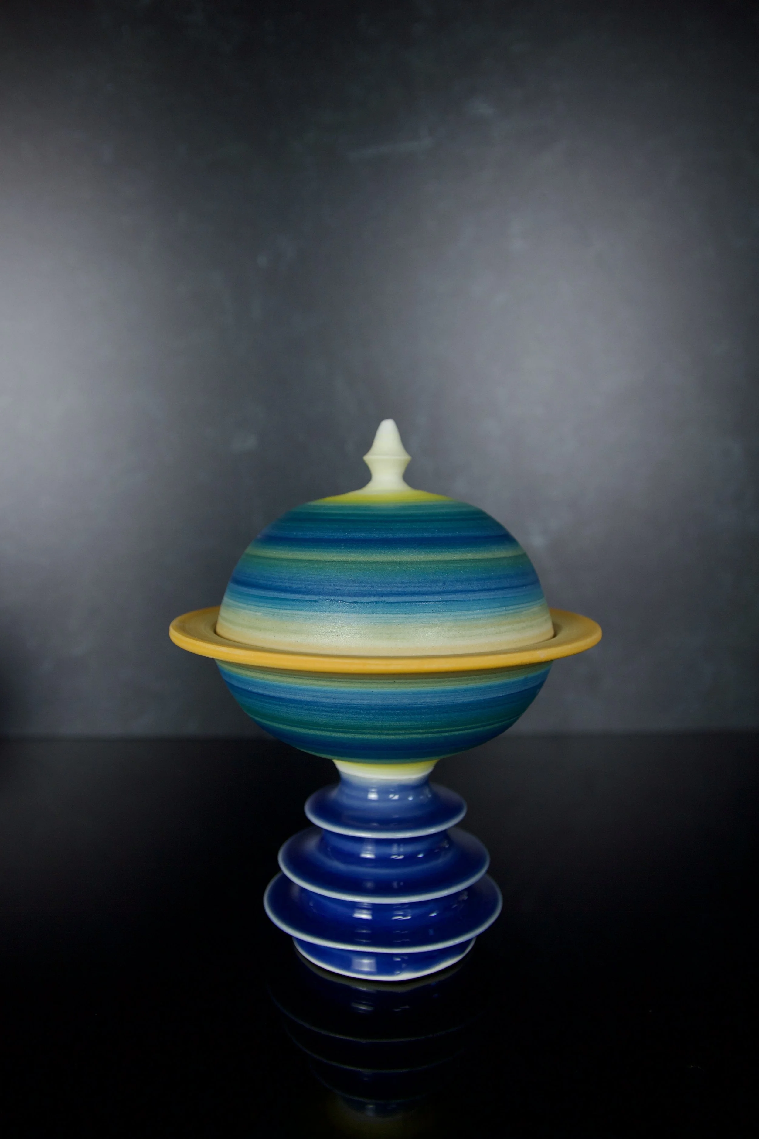 Ceramic art piece resembling a planet with rings, featuring multicolored stripes and a decorative top, displayed against a dark background.