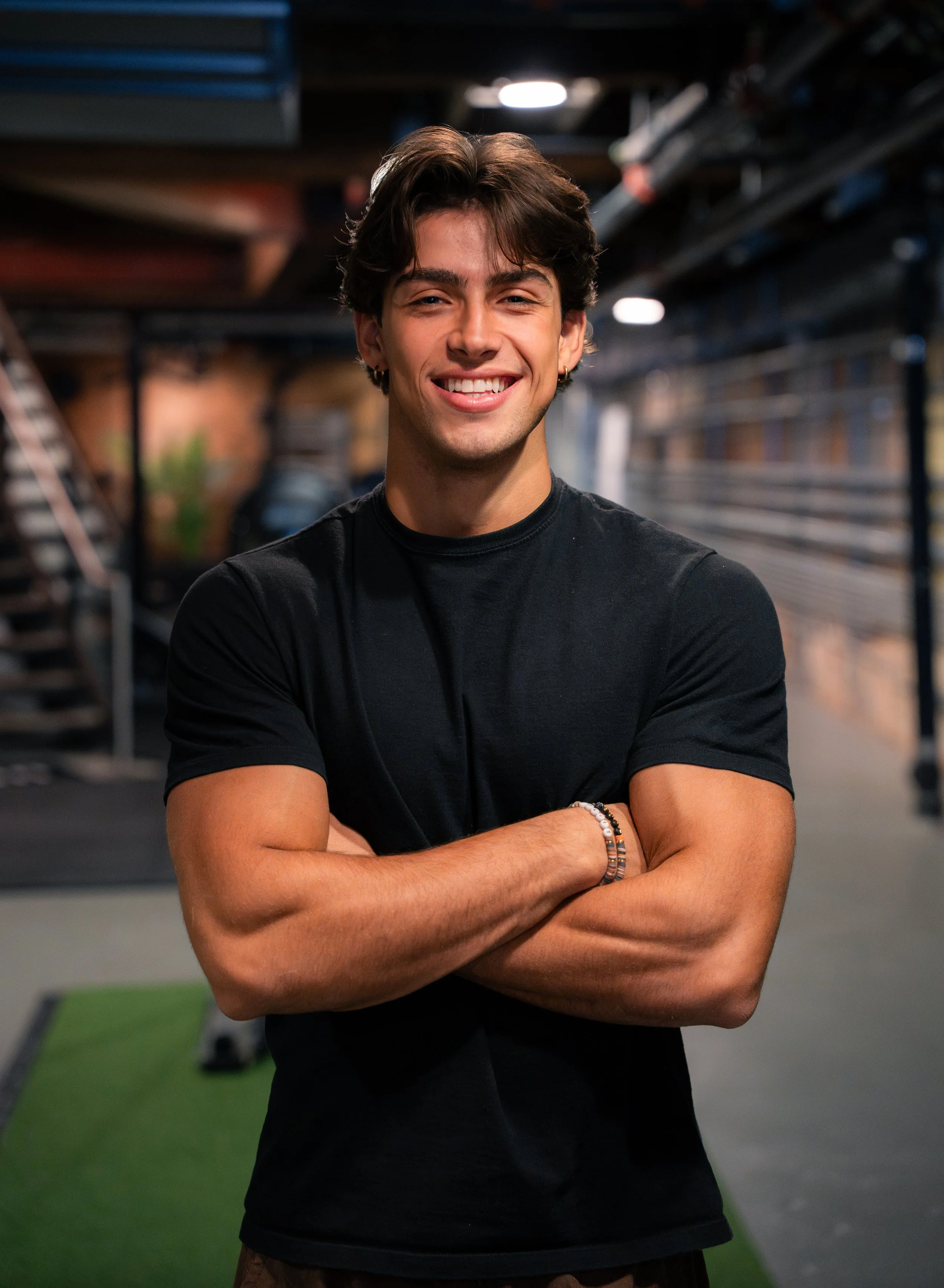 A young man with brown hair smiling at the camera, arms crossed, standing in a gym with gym equipment and stairs in the background.
