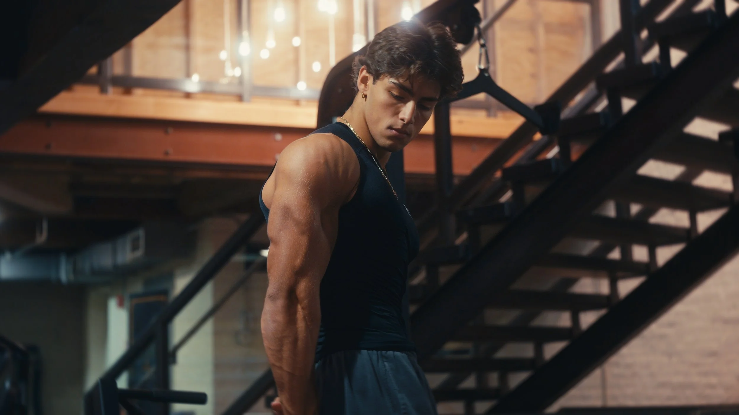 A young man with muscular arms in a black tank top and gray shorts, standing on a gym staircase indoors, looking downward, with an industrial-style interior background.