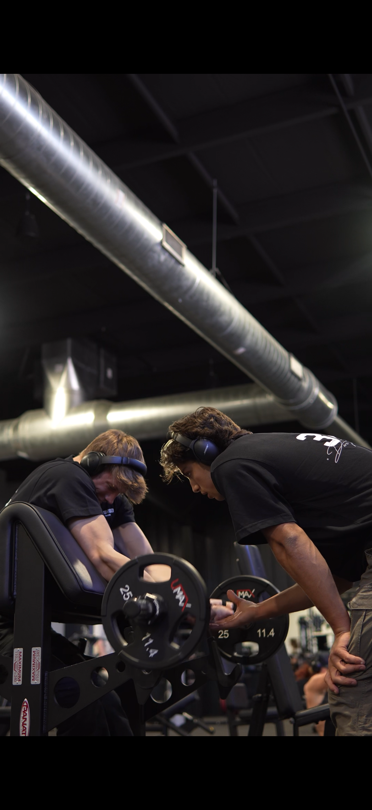 Two young men with headphones assisting an older man with stretching or warming up in a gym, with weightlifting equipment and exposed ductwork on the ceiling in the background.