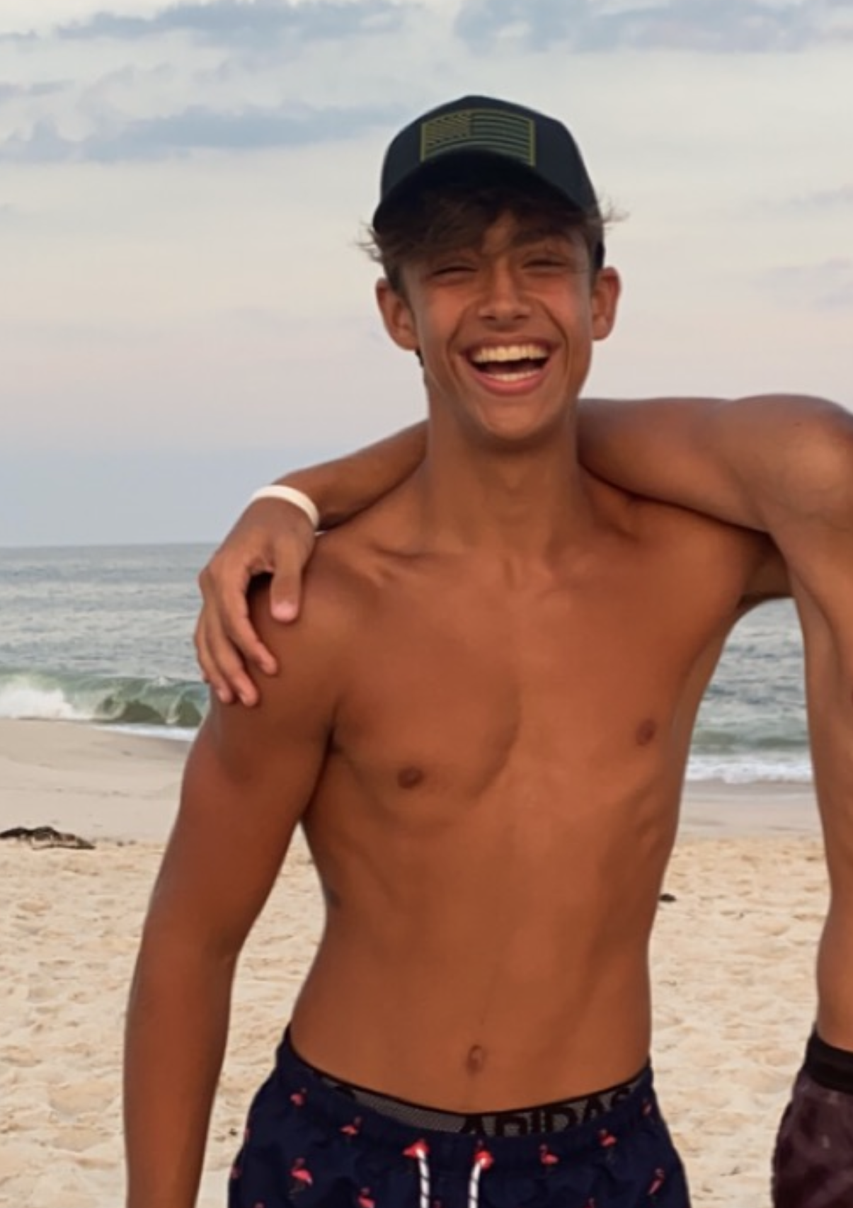A shirtless young man wearing a cap and swim trunks, smiling on a beach with the ocean and sky in the background.