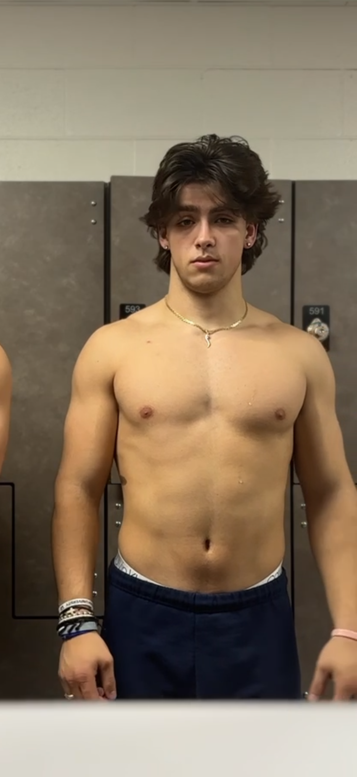 A shirtless young man with dark hair and earrings, standing in front of lockers, wearing a chain necklace and multiple bracelets.