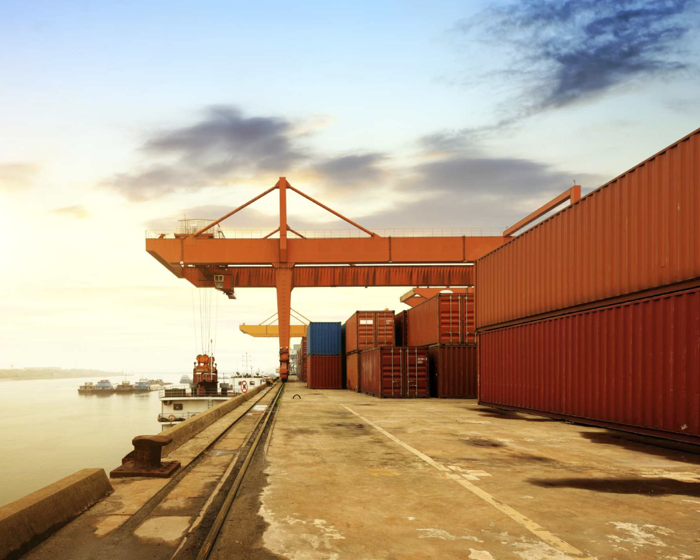 Contenedores de carga en un puerto con grúa y barcos en el fondo al atardecer.