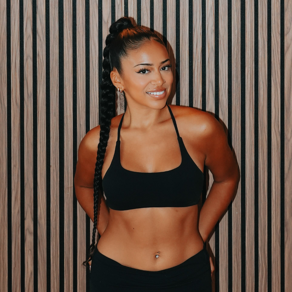 Young woman with long braided hair, wearing a black sports bra and black pants, standing against a wooden panel wall.